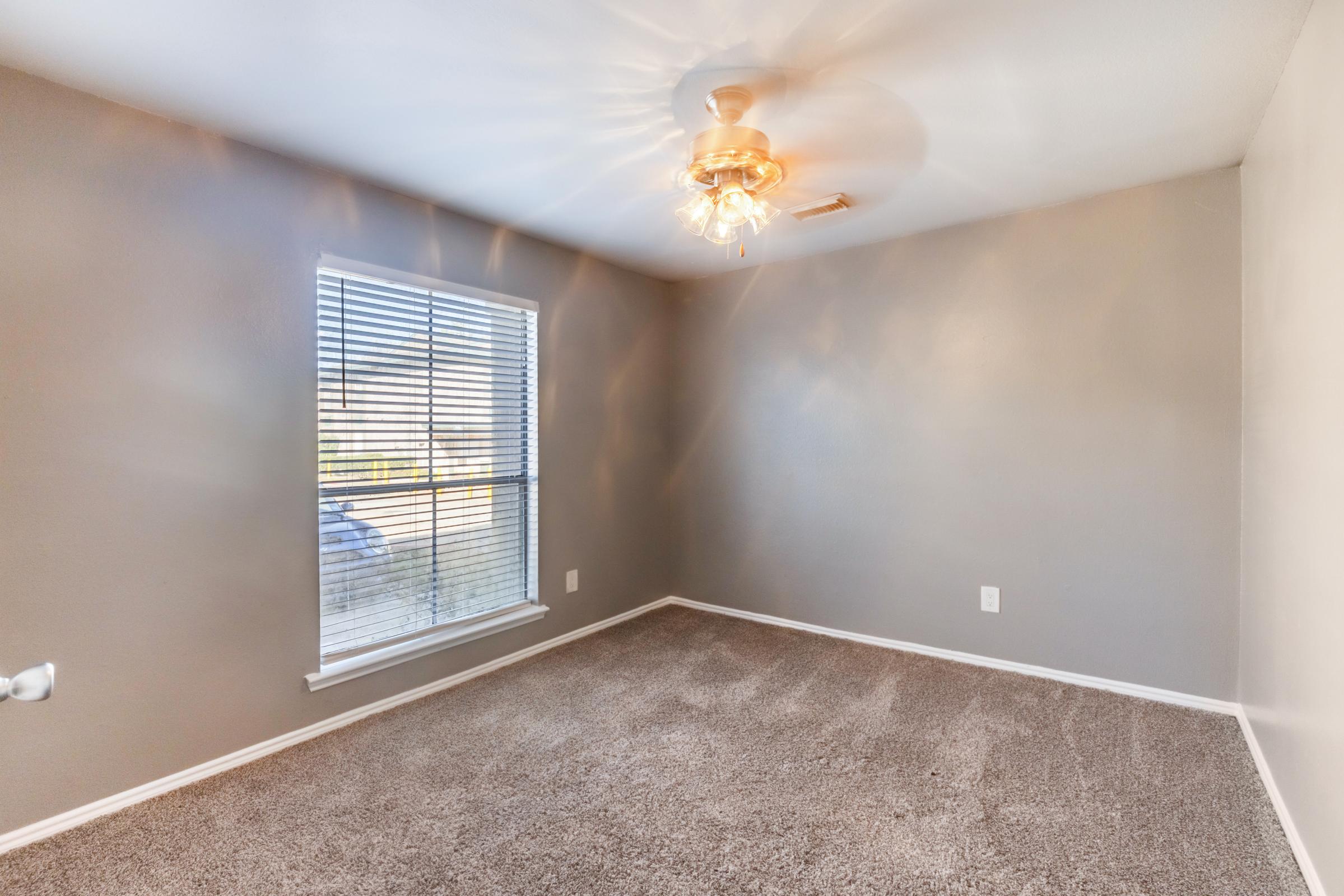 Empty room with gray walls and carpet. A ceiling fan is installed, and there is a window with blinds allowing natural light to enter. The room appears well-lit and tidy, suitable for various uses.