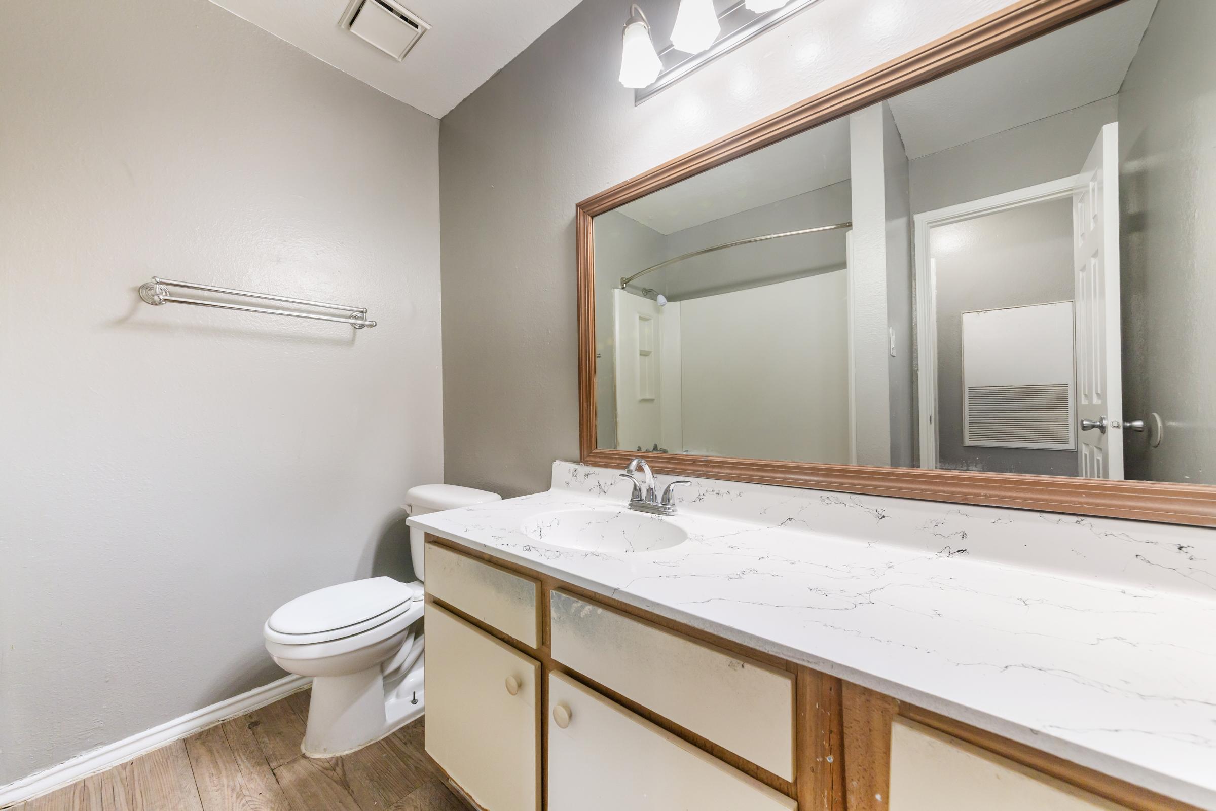 A modern bathroom featuring a white toilet, a sink with a marble countertop, and a large mirror above the sink. The walls are painted gray, and there is a towel rack on the side. A door leading to another room is visible, along with light fixtures above the mirror. The flooring appears to be a brown wood-like material.