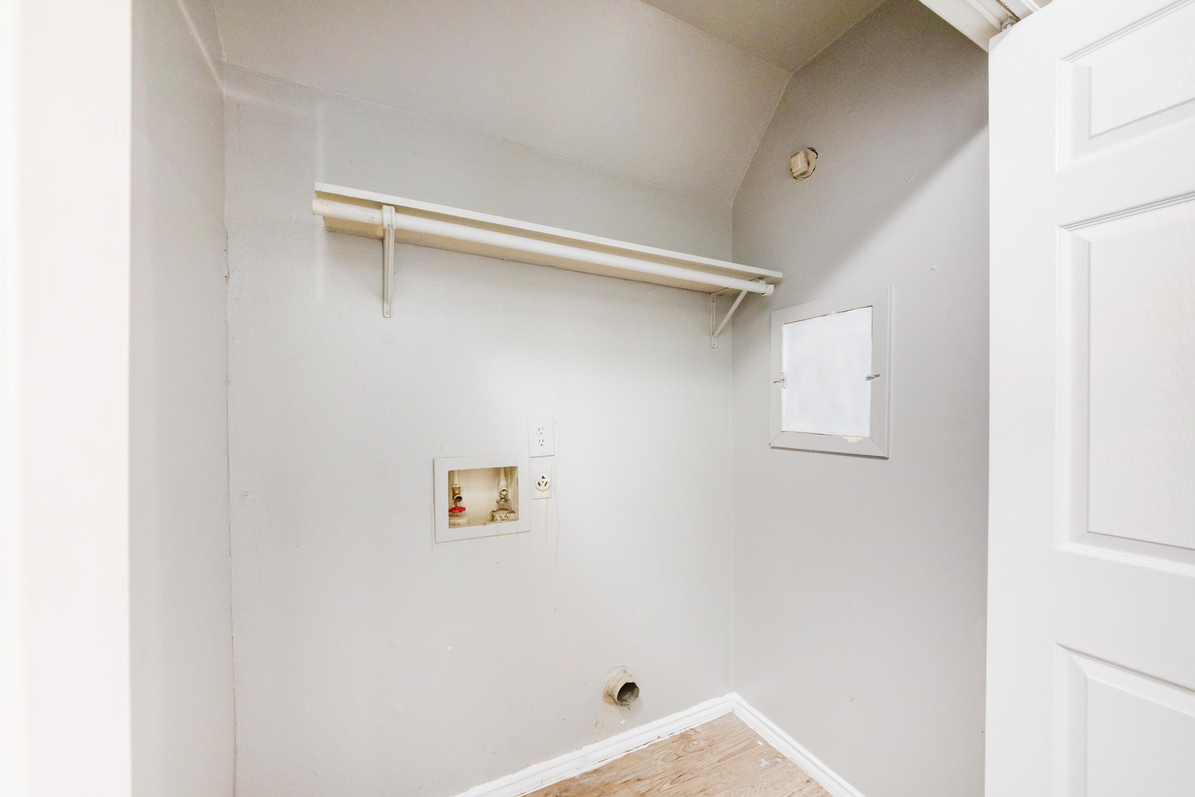 A small, empty room with light gray walls and a sloped ceiling. There is a single shelf mounted on the wall and a small window or cutout on one side. The floor is bare, and a circular hole is visible in the bottom wall, indicating a possible utility access point. The door frame is visible on the right.