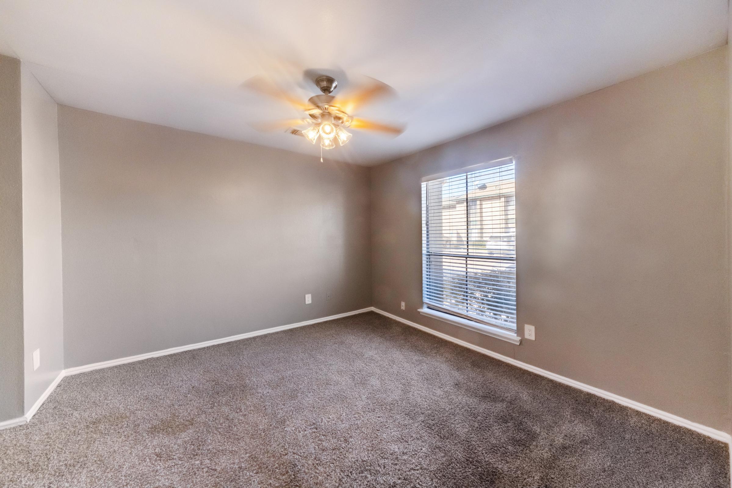 A spacious, empty room with light gray walls and carpeted flooring. A ceiling fan with four lights is mounted in the center, and a large window with closed blinds allows natural light to enter the room. There are no furnishings, creating a bright and open atmosphere.