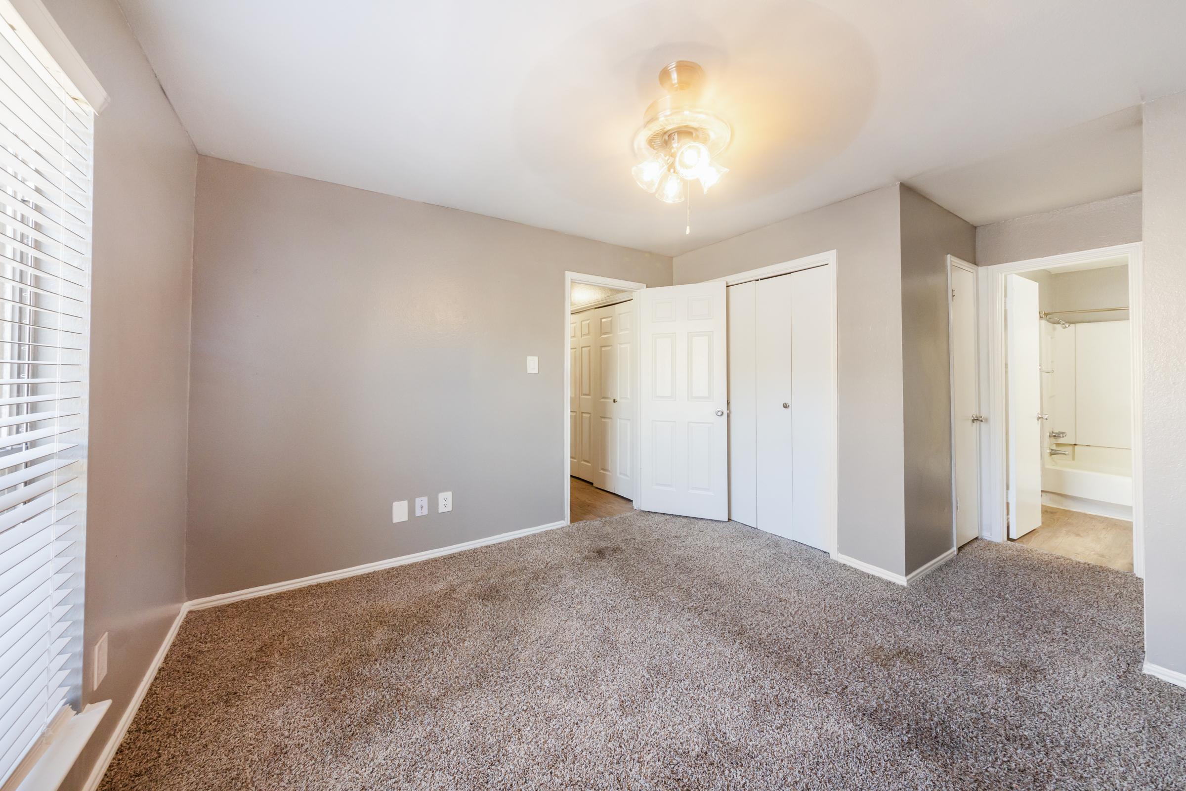 A spacious, well-lit room with grey walls and carpet flooring. It features a ceiling fan, two closet doors, and an adjoining bathroom visible through an open door. Natural light streams in from a window with blinds. The overall ambiance is fresh and inviting, suitable for various uses.