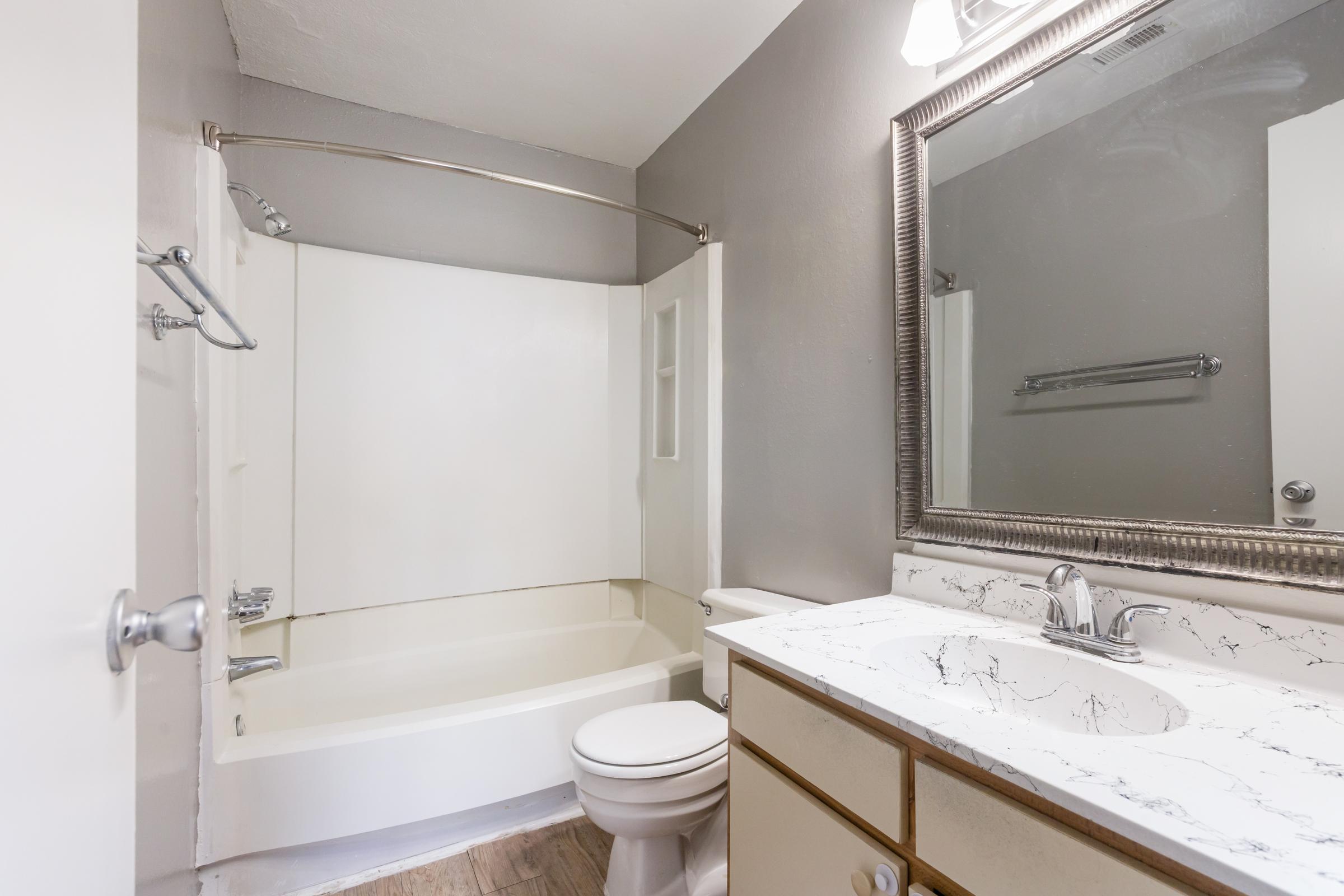A clean and modern bathroom featuring a shower/tub combo, a white vanity with a marble countertop, a mirror above the sink, and light gray walls. The space is well-lit and organized, with a toilet and a towel rack on the wall.