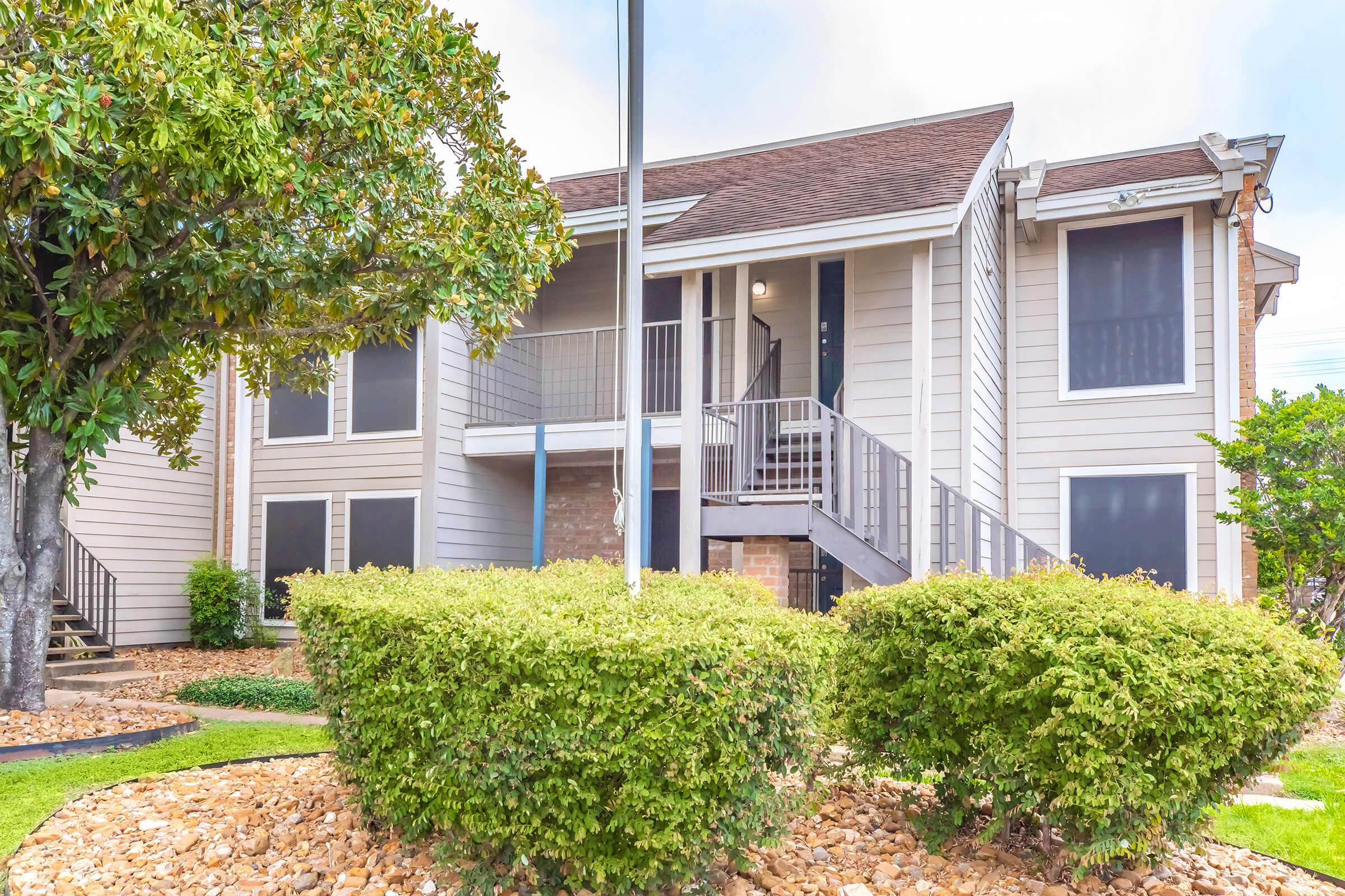 A two-story apartment building featuring a staircase and covered balcony, surrounded by well-maintained landscaping with shrubs and gravel. A flagpole is positioned in the front yard area, contributing to the welcoming appearance of the property.