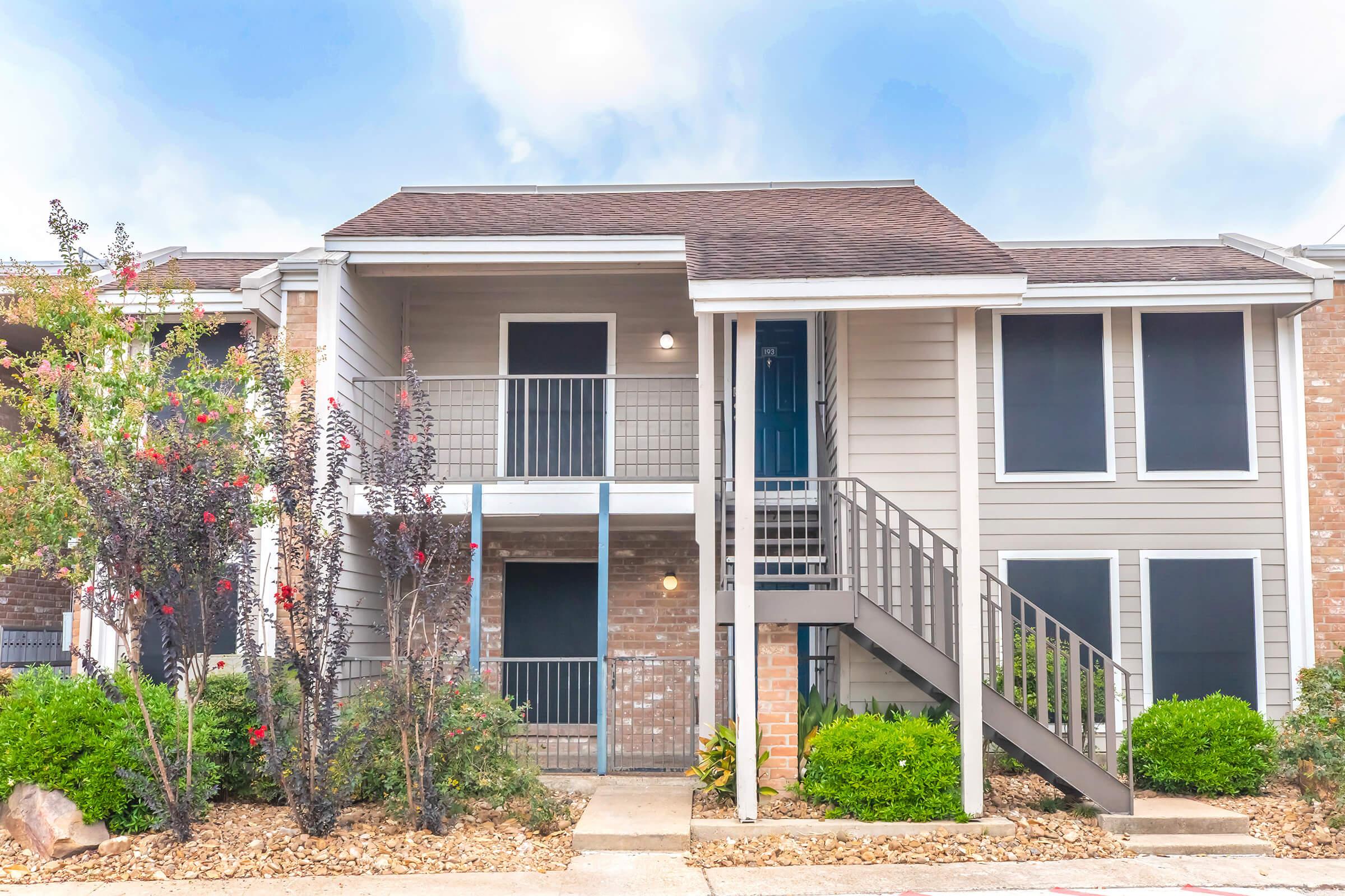 A two-story apartment building with a light-colored exterior and brick accents. The entrance features a staircase leading to the upper level. Surrounding the building are small shrubs and decorative plants, and the scene is bright with a partly cloudy sky.