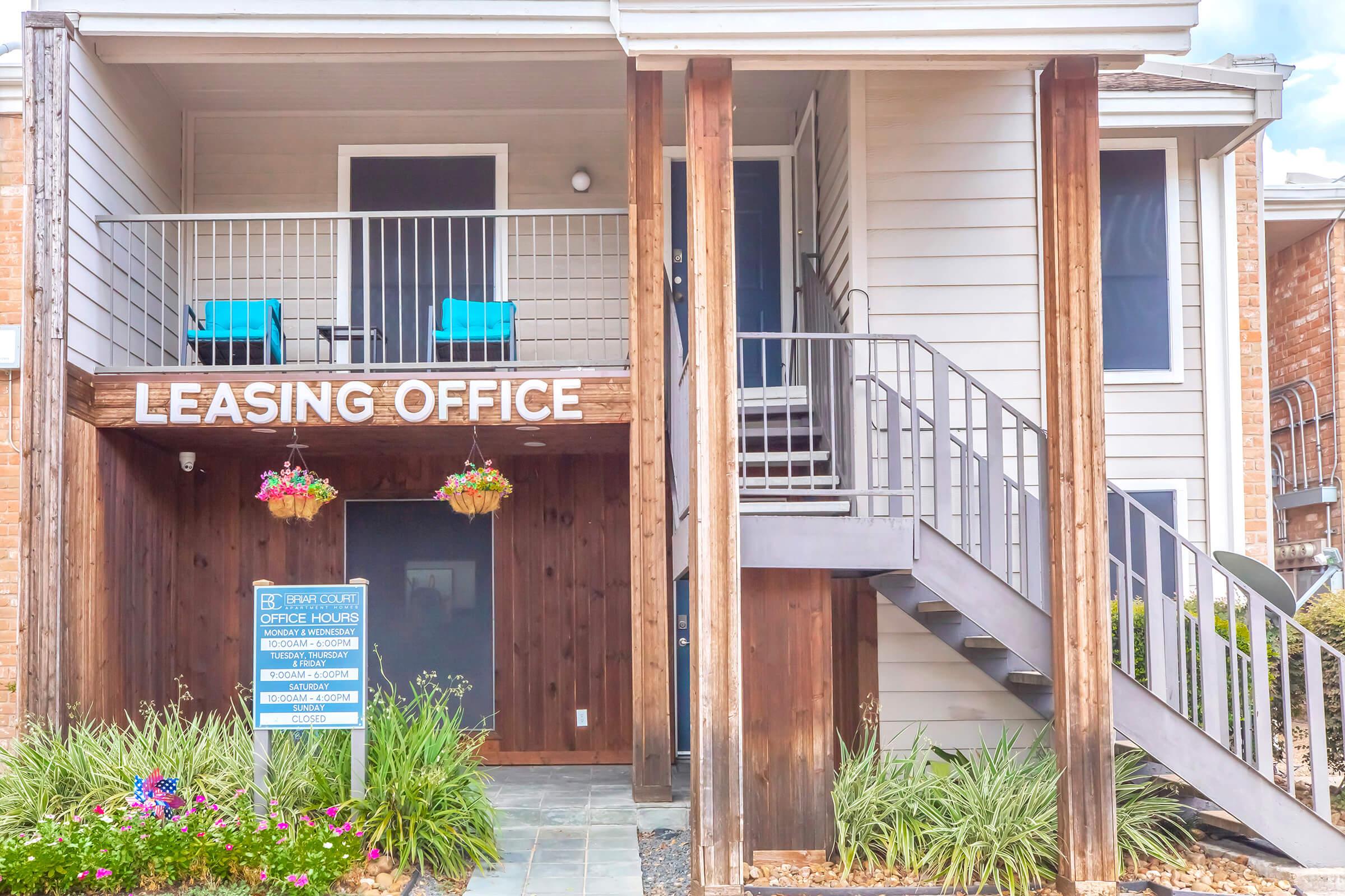 A leasing office entrance featuring a wooden sign that reads "LEASING OFFICE." The building has two blue chairs on the porch, a stairway leading to the second floor, and decorative hanging plants. A sign on the ground details office hours and contact information, surrounded by green shrubs and landscaping.