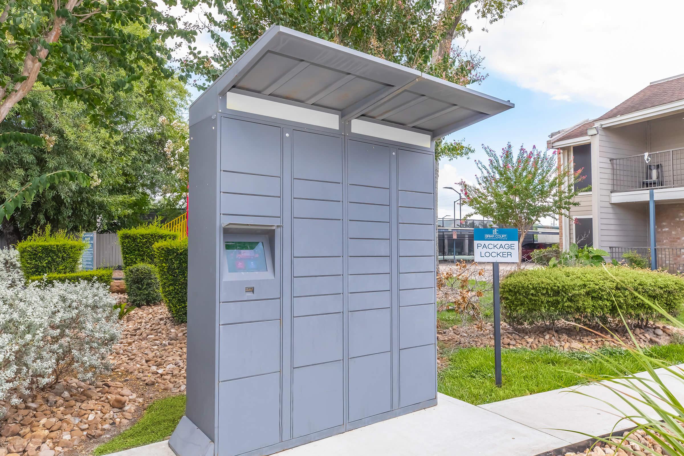 A gray package locker unit with a roof, located outdoors in a landscaped area. The locker has a digital interface on one side, surrounded by greenery and decorative plants. A sign nearby indicates it is a package locker for deliveries.