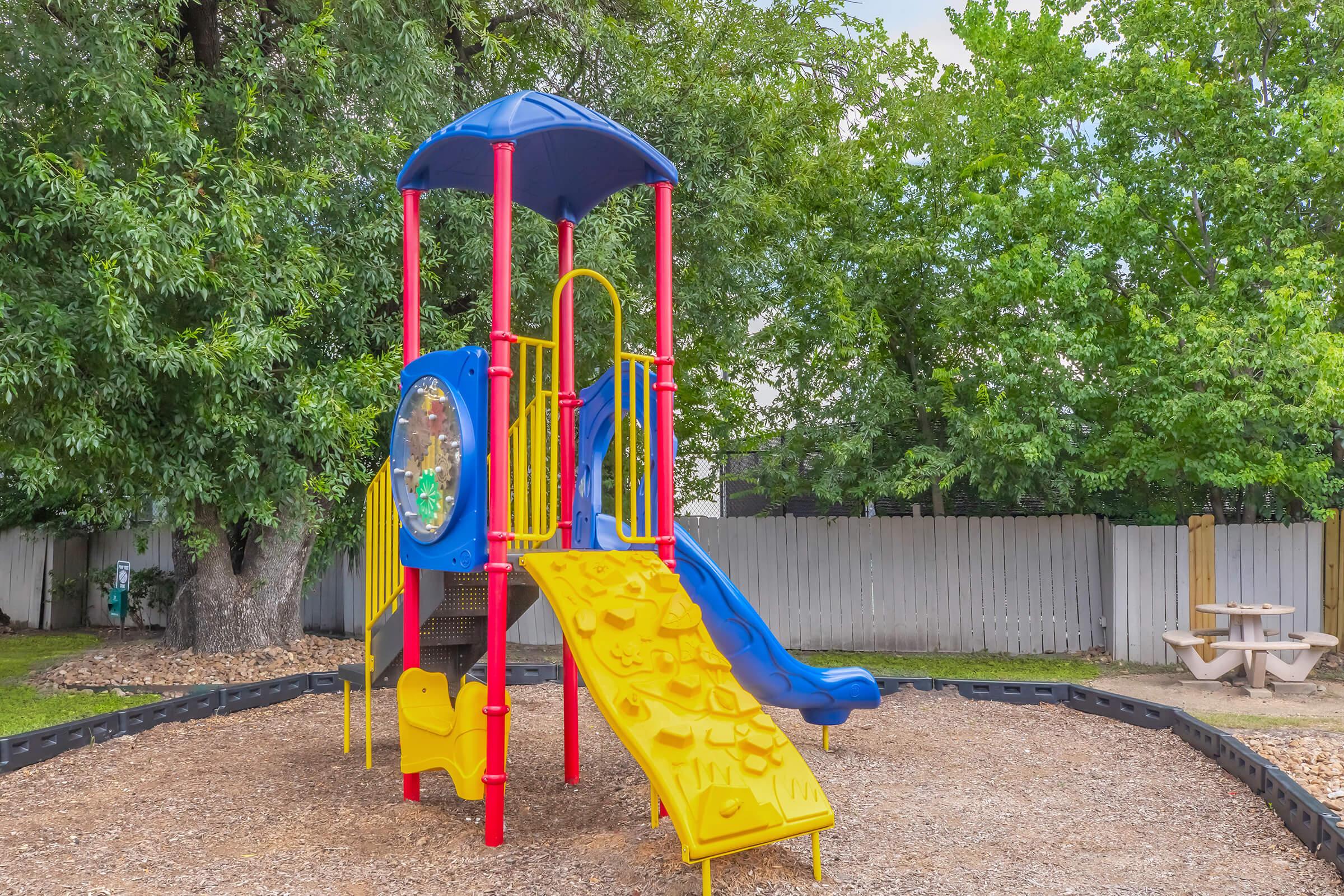 Colorful playground equipment featuring a blue and red structure with a slide, climbing wall, and an observation window. The playground is surrounded by greenery, with a gravel base and a picnic area in the background. Ideal for children to enjoy outdoor play.