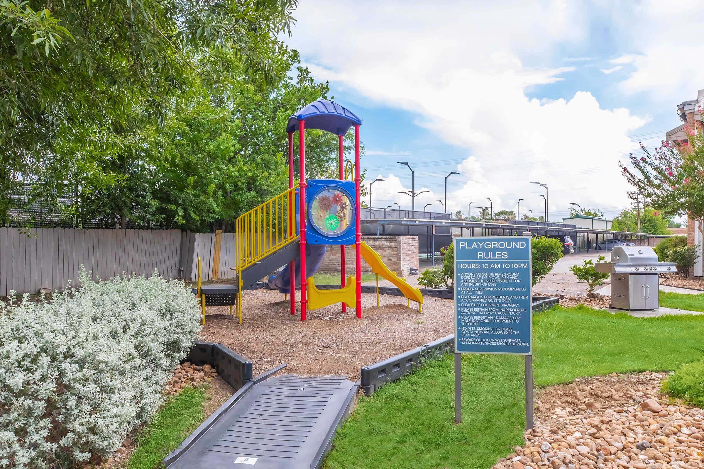 A colorful playground structure featuring a slide and climbing area, surrounded by green grass and landscaping. A sign nearby provides information about playground rules and hours. Bright blue skies with scattered clouds are visible in the background.