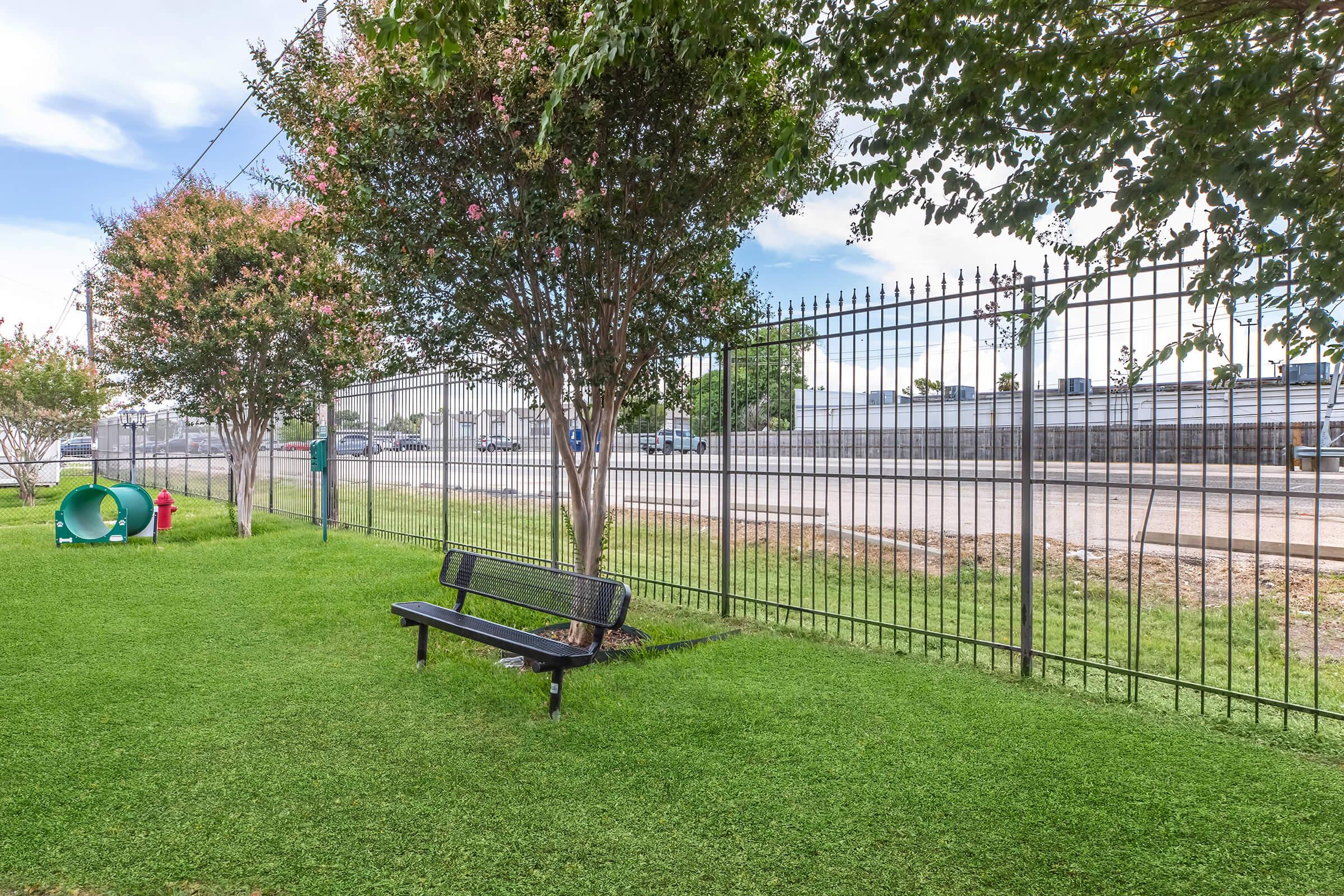 A grassy area with a black bench underneath a tree, bordered by a black metal fence. In the background, a road with parked vehicles is visible. A small green playground structure is located on the left side of the image. The sky is partly cloudy.