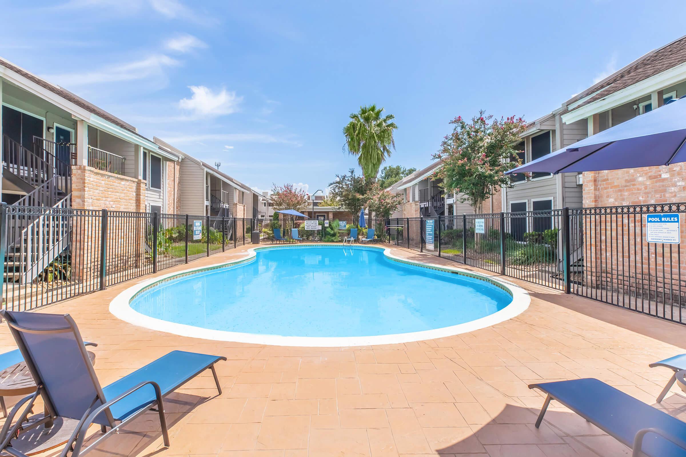 A well-maintained outdoor swimming pool surrounded by lounge chairs, with palm trees and flowering plants in the background. The pool area is enclosed by a black fence, and there are two-story apartment buildings visible in the background under a clear blue sky.