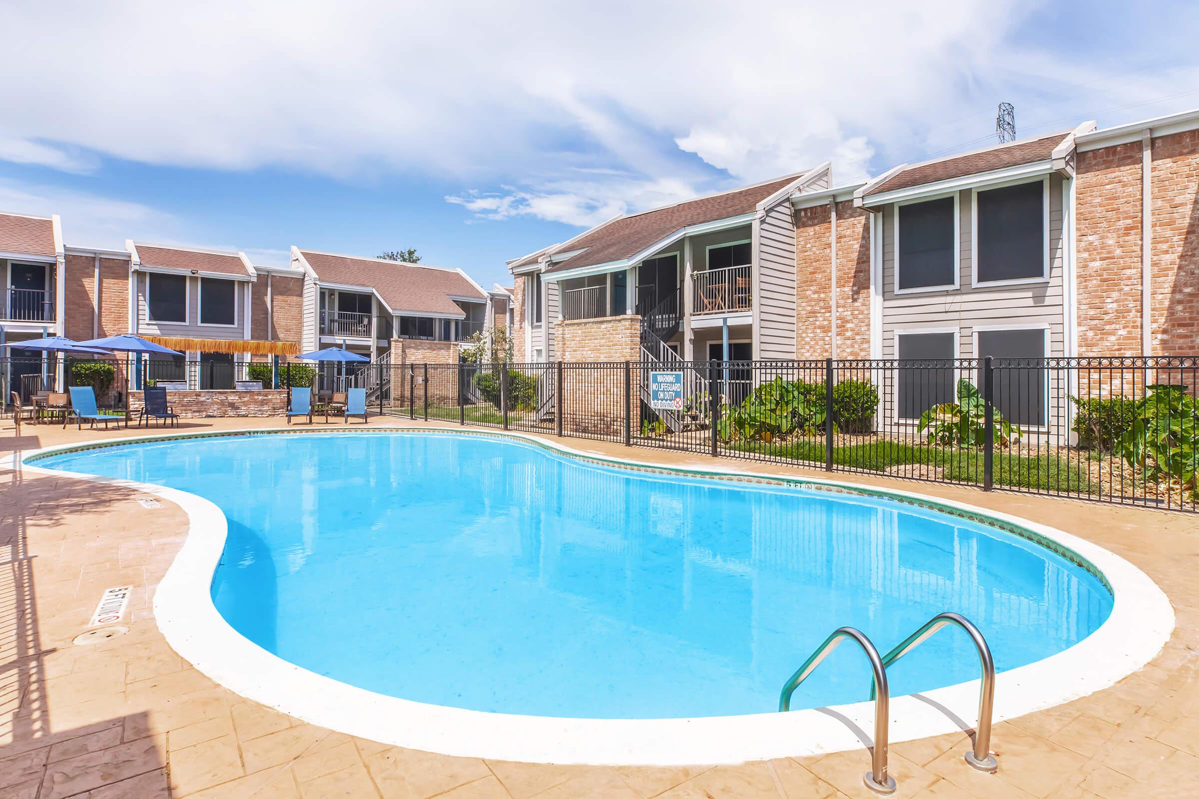 A clear blue swimming pool surrounded by a pool deck and lounge chairs. In the background, there are several residential buildings with brick exteriors and screened windows. The sky is bright and partly cloudy, creating a sunny atmosphere. A fence encloses the pool area.