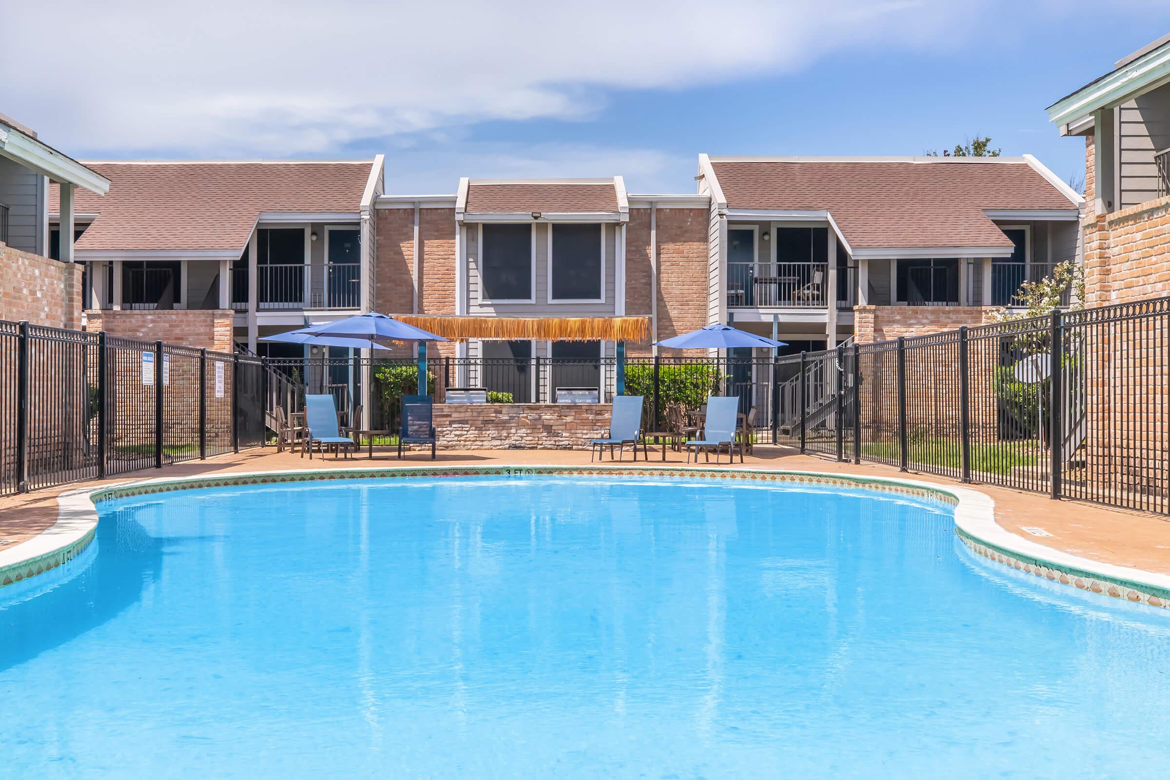 A swimming pool surrounded by a fenced area, with lounge chairs and umbrellas set up on the pool deck. In the background, there are apartment buildings with balconies. The sky is clear with a few clouds, creating a bright and inviting atmosphere.