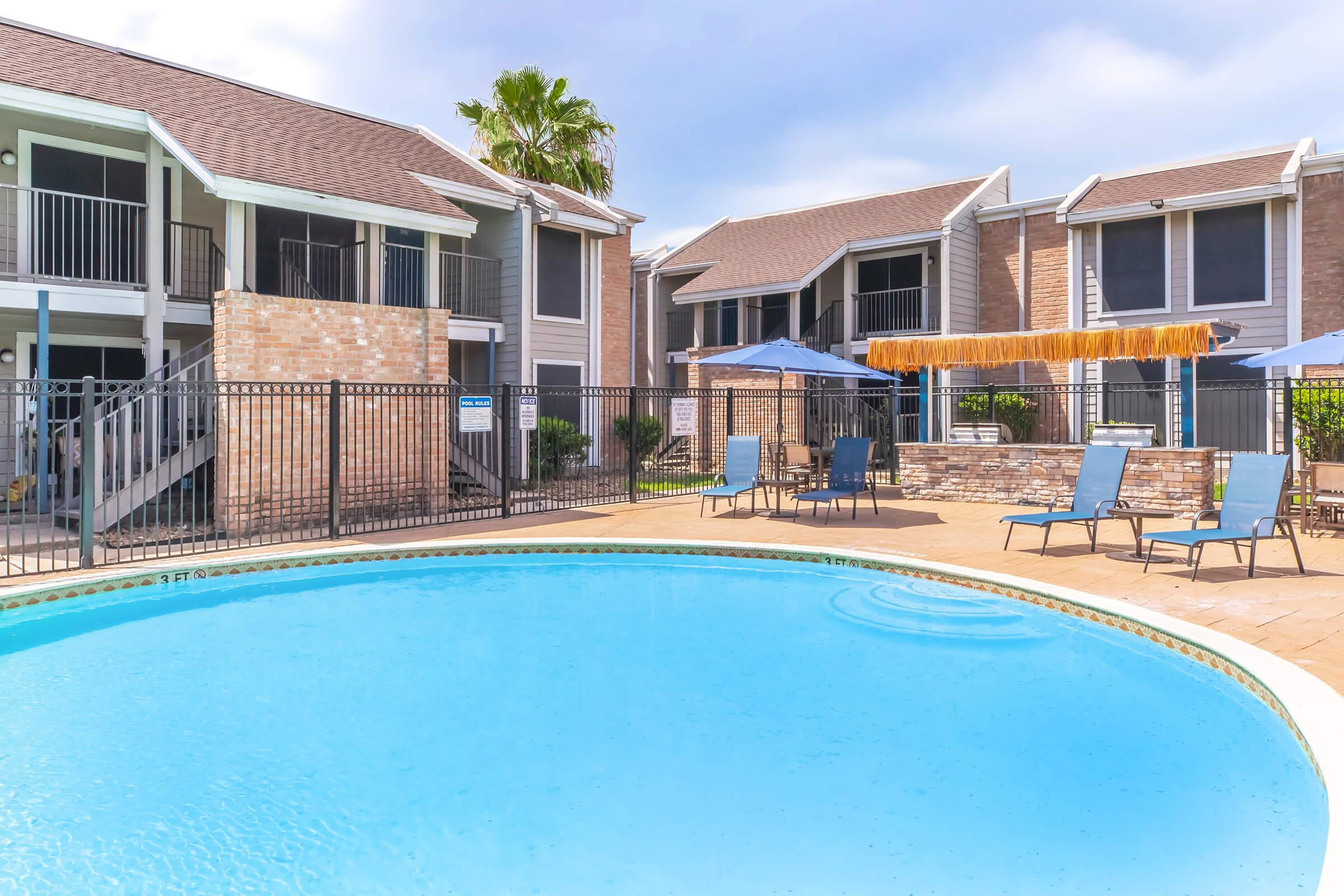 A swimming pool surrounded by a sun patio featuring lounge chairs and umbrellas. In the background, two multi-story apartment buildings with balconies are visible. The area is well-lit with palm trees, creating a relaxing outdoor atmosphere.