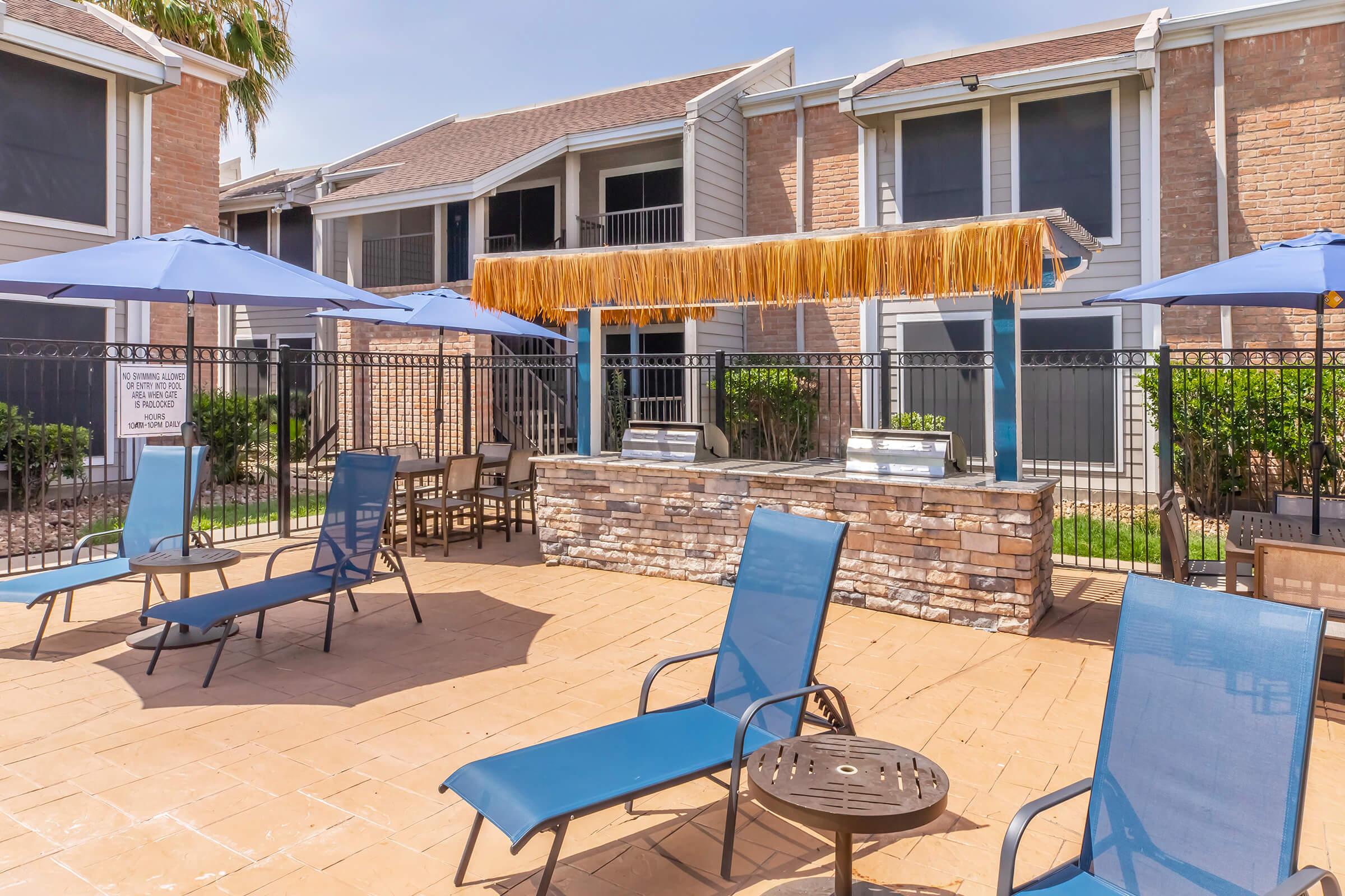 A sunny pool area featuring blue lounge chairs, umbrellas, and a thatched-roof bar. The surrounding buildings are residential, with greenery and a fence in the backdrop. The atmosphere is relaxed and inviting, perfect for leisure and socializing by the pool.