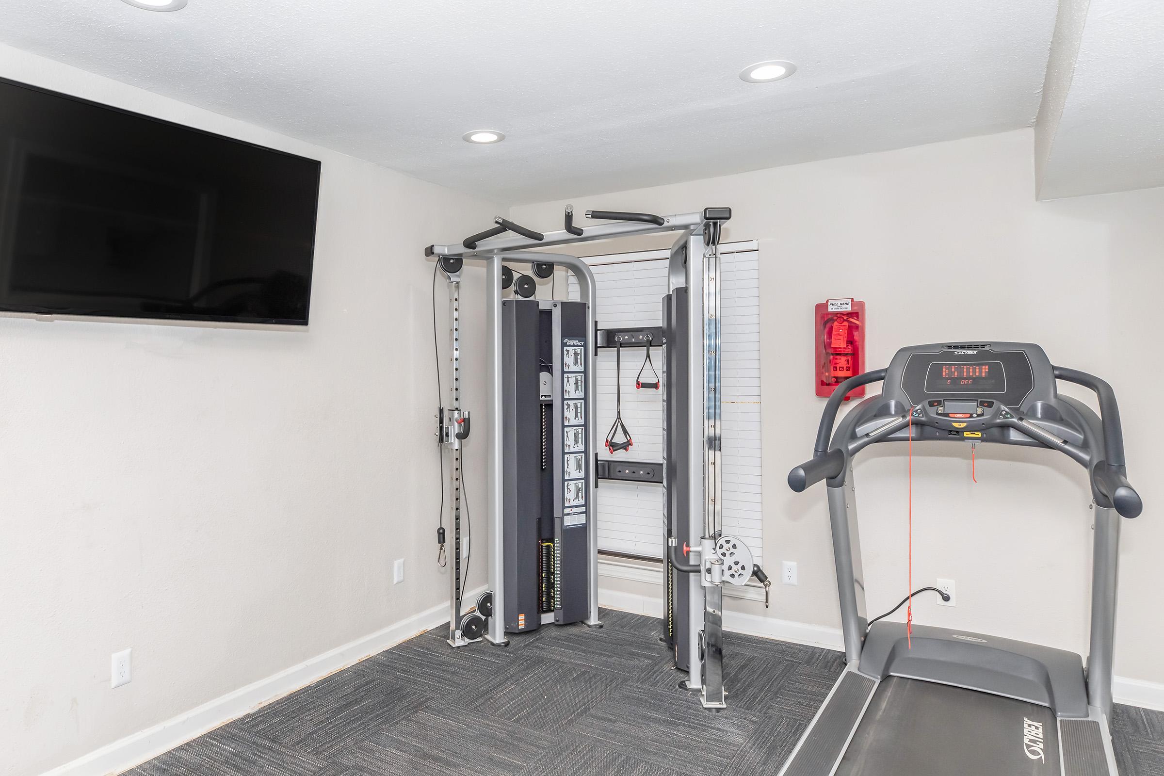 A modern home gym featuring a multi-station workout machine, a treadmill, and a wall-mounted TV. The room has light-colored walls, a window with blinds, and a fire extinguisher visible for safety. The floor is covered with dark carpet, providing a clean and functional workout space.