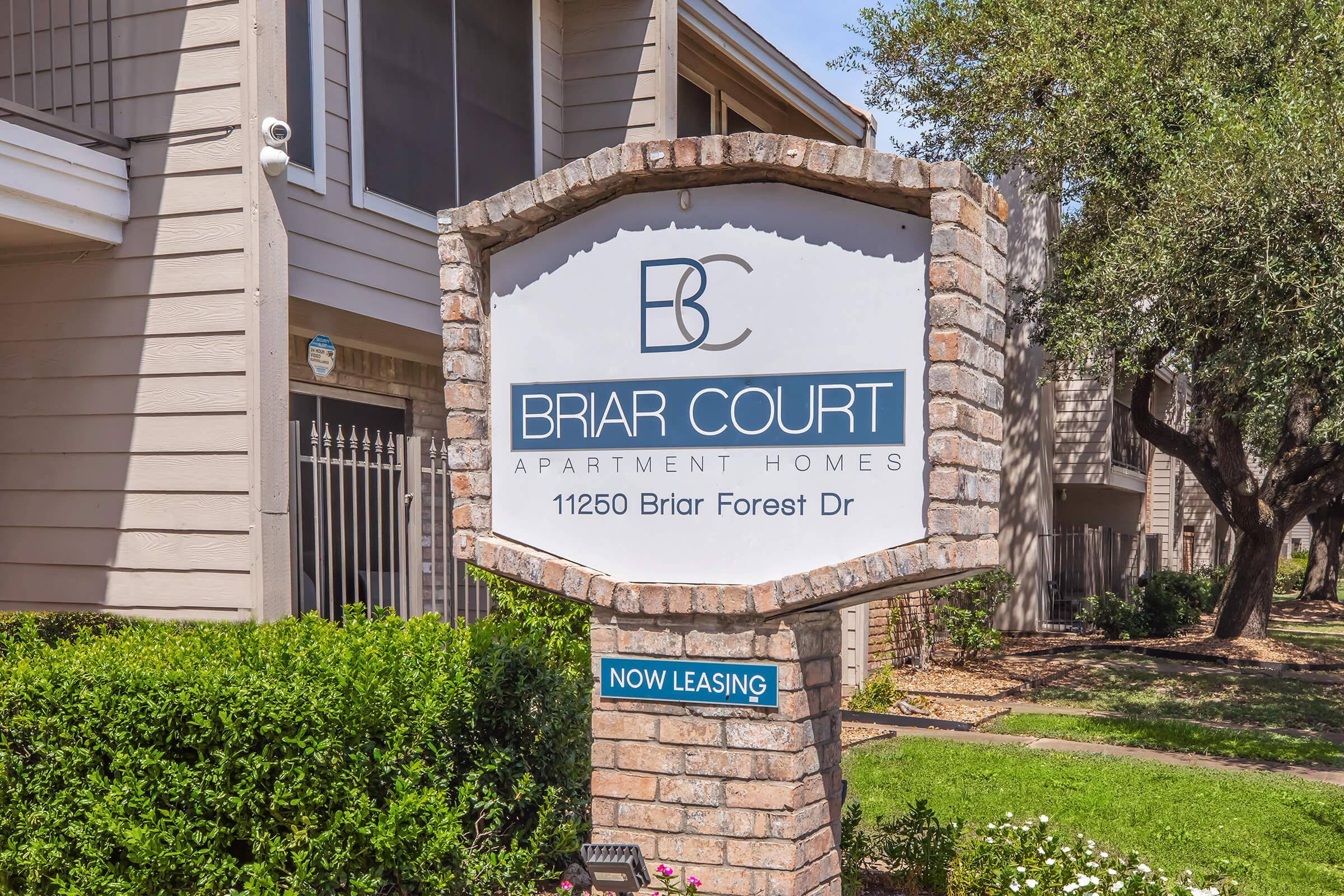 Sign for Briar Court Apartment Homes, located at 11250 Briar Forest Dr. The sign features a brick border and indicates that the apartments are currently leasing, with a "Now Leasing" label prominently displayed below the name. Surrounding greenery and trees are visible in the background.