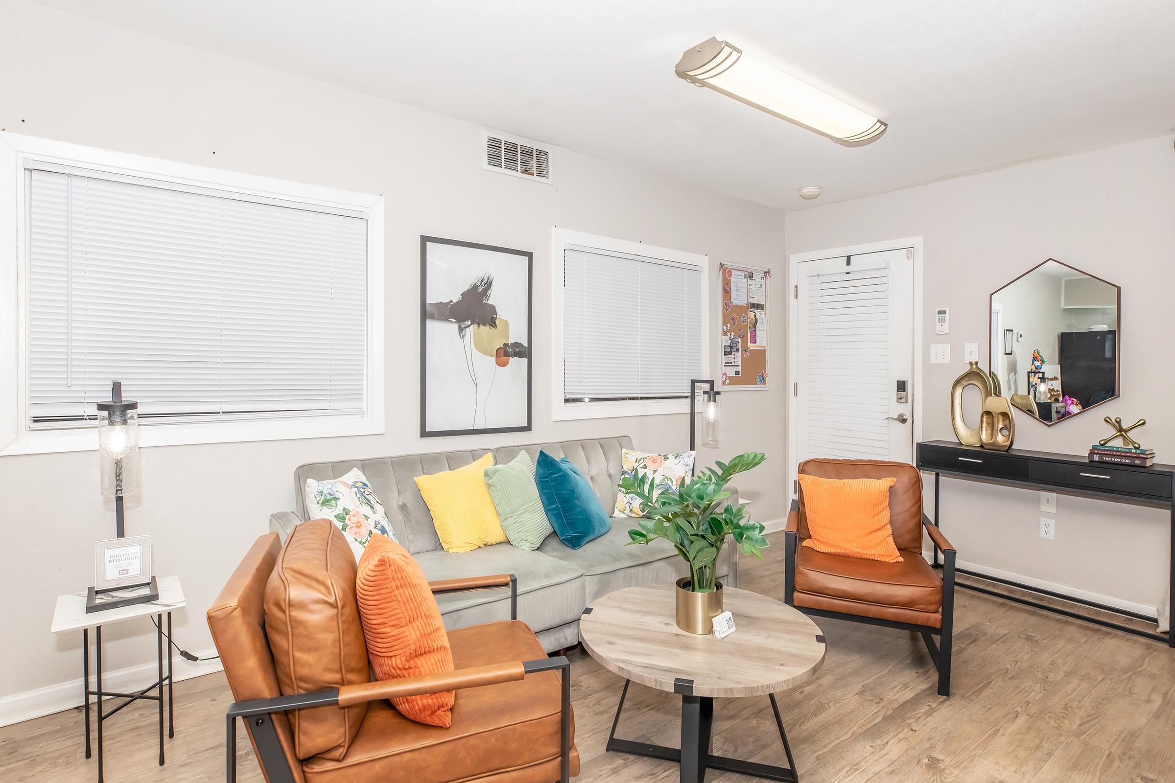 A cozy living room featuring a gray sofa with colorful cushions, two orange armchairs, a round wooden coffee table with a plant, and a stylish black console table. Natural light streams in through white blinds, and there's a bulletin board on the wall, creating a welcoming atmosphere.