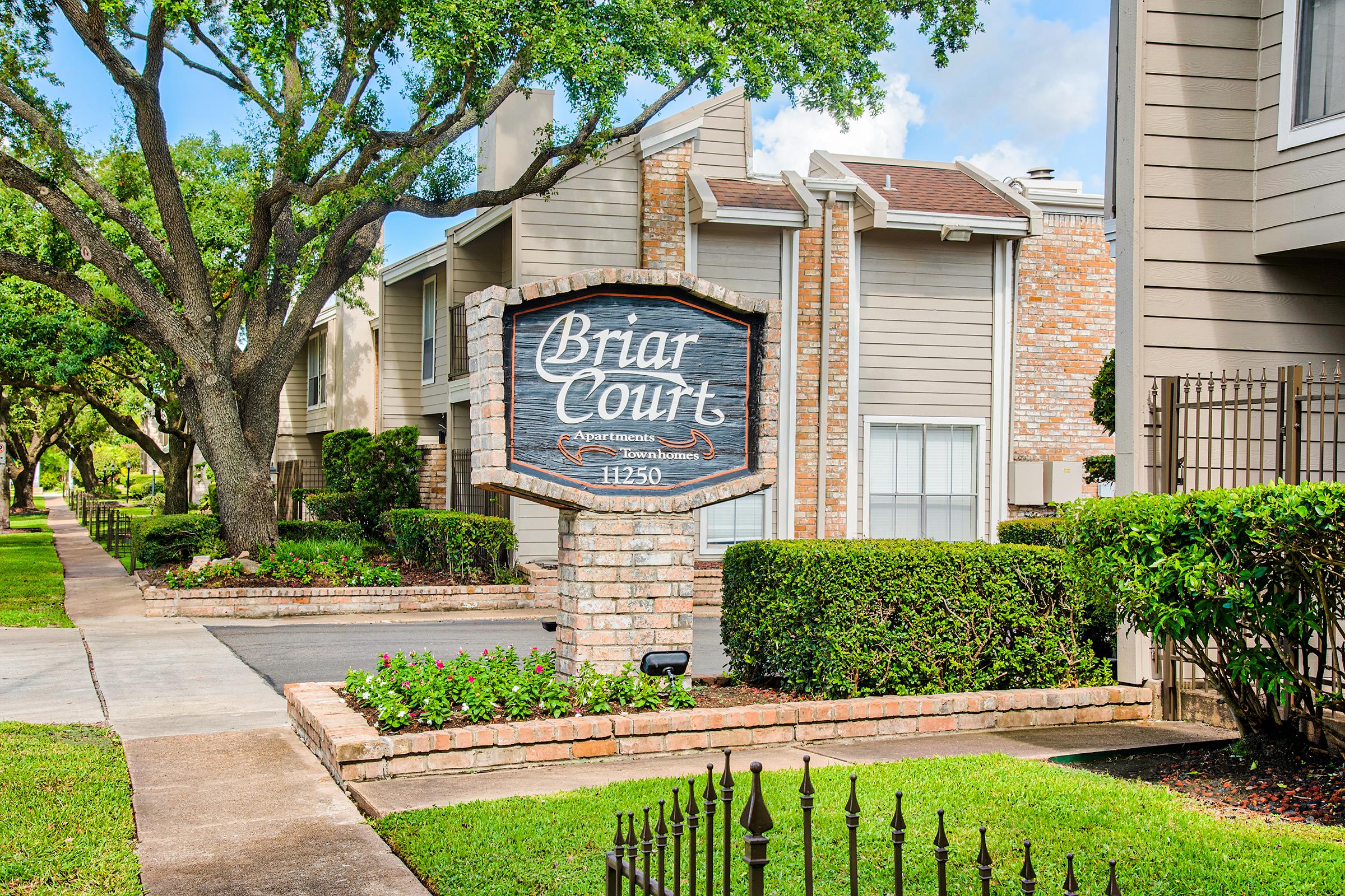 Sign for Briar Courts Apartments with stone and wooden elements, surrounded by well-maintained landscaping, including bushes and flowers. There are trees lining the walkway, with residential buildings visible in the background under a partly cloudy sky.
