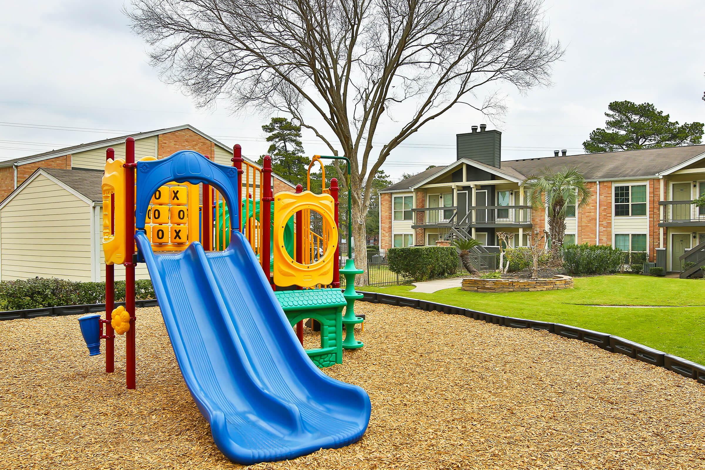 A colorful playground featuring a blue slide and a green play structure, set on a bed of wood chips. In the background, there are residential apartment buildings surrounded by grass and trees, with overcast skies above.