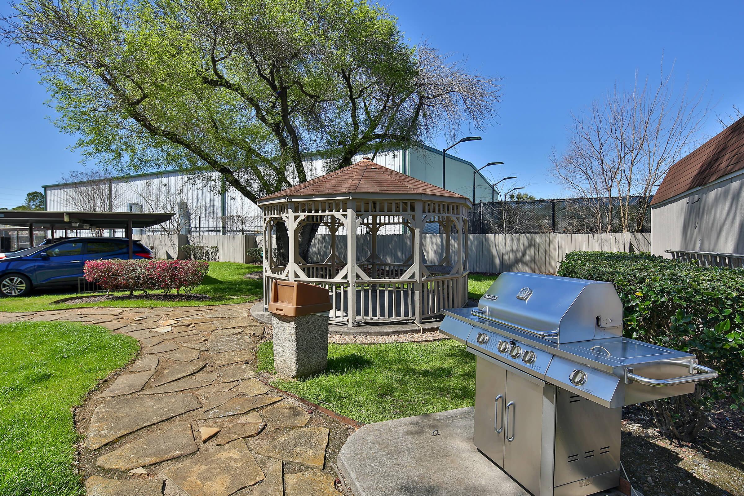 A well-maintained outdoor area featuring a gazebo, a stone pathway, a barbecue grill, and landscaped greenery. In the background, there are trees and a building, creating a pleasant and inviting atmosphere suitable for gatherings and relaxation.