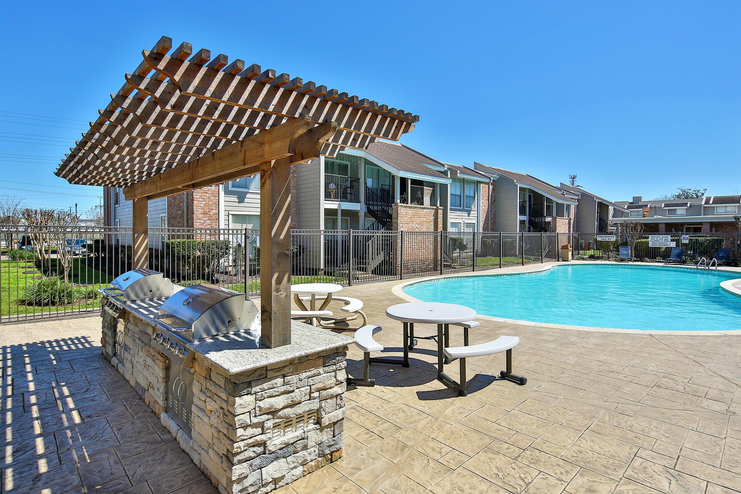 An outdoor area featuring a swimming pool surrounded by a fence, with a pergola that includes barbecue grills and picnic tables. The scene is bright and sunny, showcasing buildings in the background and a well-maintained lawn. Ideal for social gatherings and relaxation.