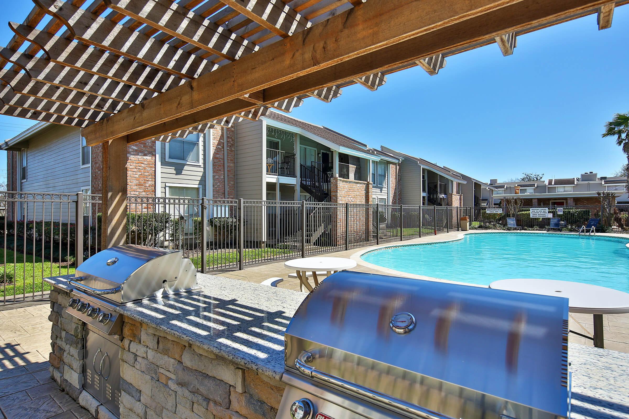 A sunny outdoor area featuring a swimming pool and a shaded barbecue grill under a wooden pergola. Surrounding the pool are several apartment buildings, and there's a fence for safety. Tables are arranged near the pool for relaxation and dining. The scene conveys a pleasant, inviting atmosphere.