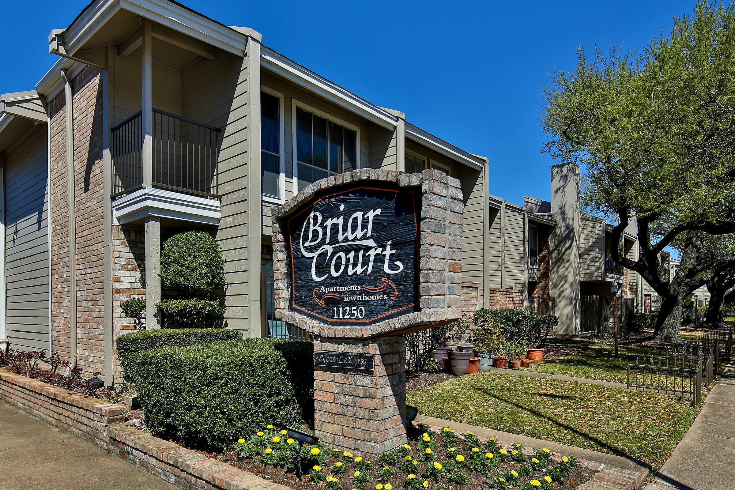 Sign for Briar Court Apartments, featuring the name prominently displayed. The scene shows well-maintained apartment buildings with a landscaped area, including shrubs and flowers, under a clear blue sky. The structure of the apartments appears modern and inviting.