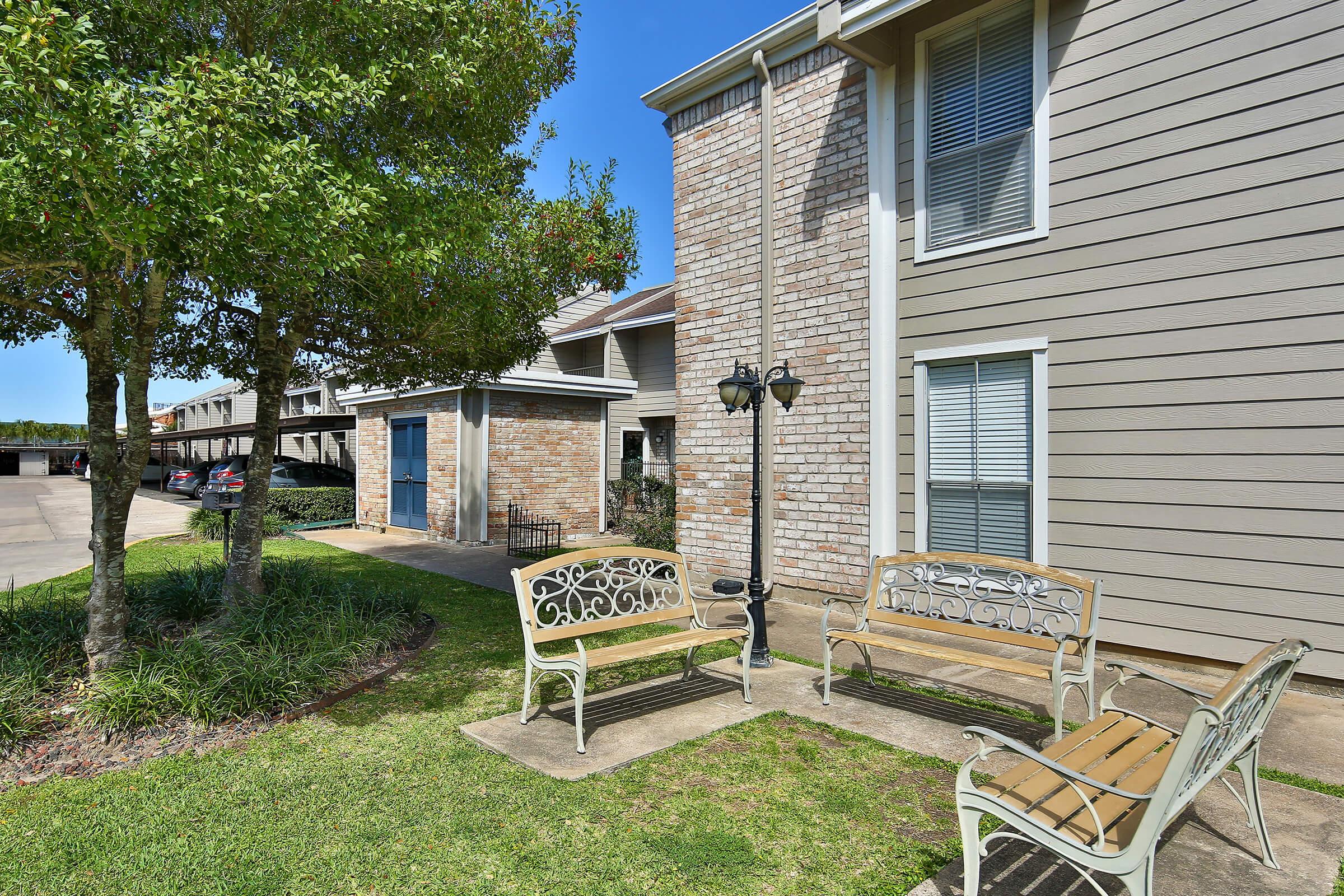 A well-maintained outdoor area featuring two decorative benches, surrounded by lush greenery and trees. In the background, there's a building with a light brick facade and a blue door, along with parked cars visible in a nearby parking lot under clear blue skies.
