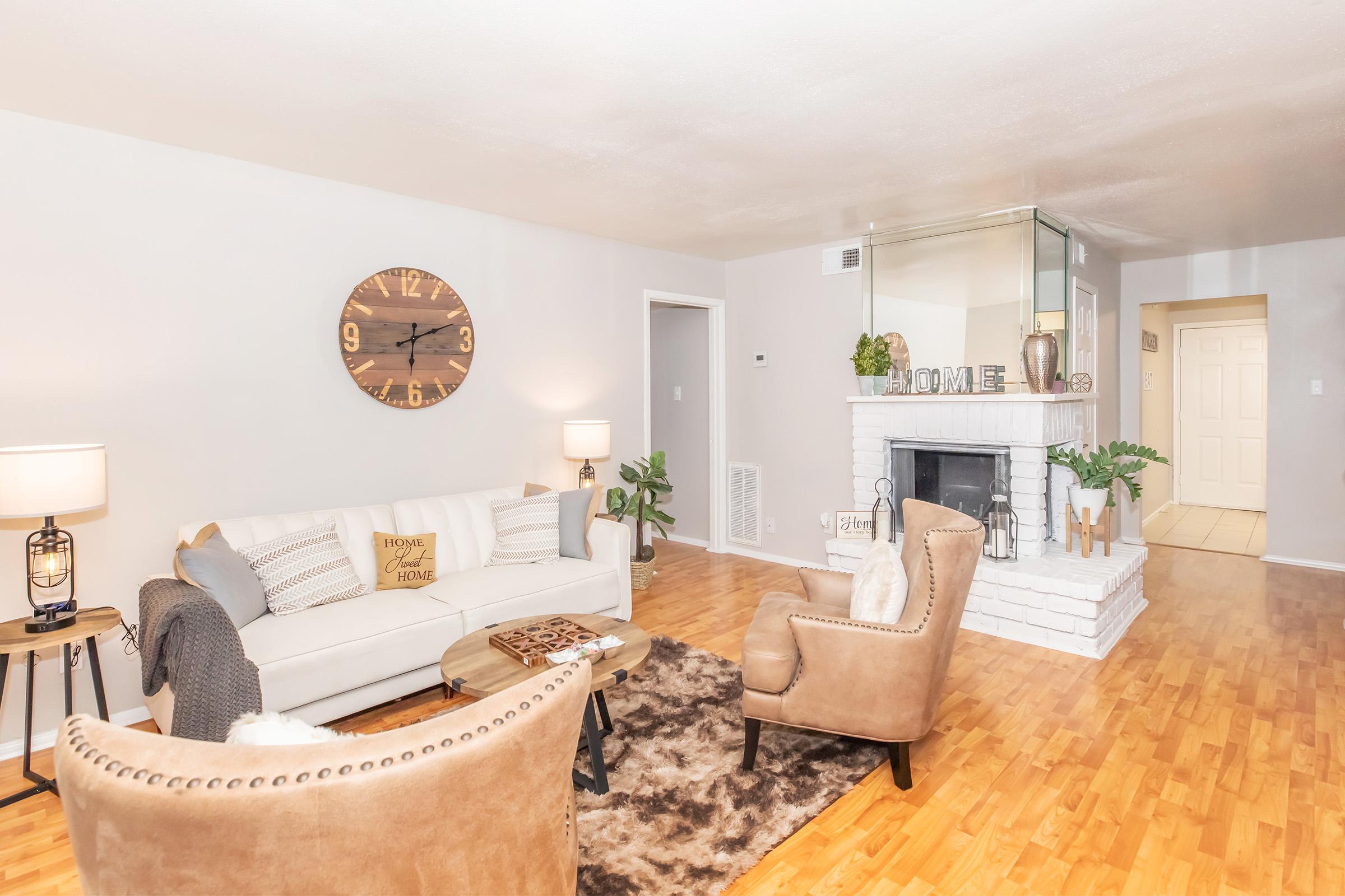 Cozy living room featuring a light-colored sofa adorned with decorative pillows, a round wooden clock on the wall, and a plush area rug. Two upholstered chairs sit beside a small coffee table. The space is illuminated by warm lighting, with a fireplace and a minimalist decor reflecting a welcoming atmosphere.