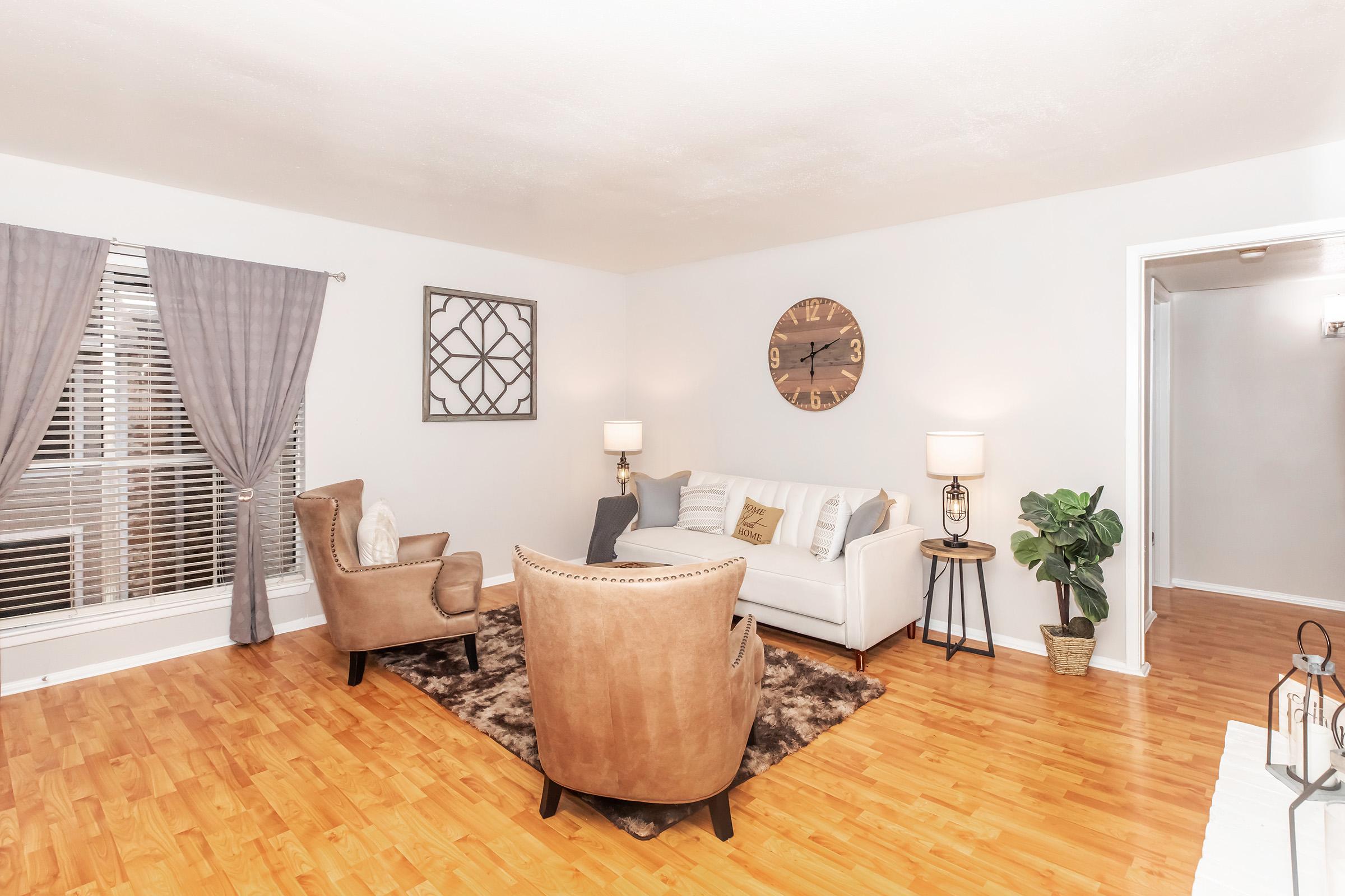 A cozy living room featuring a white couch with decorative pillows, two brown armchairs, and a textured area rug. Large windows with gray curtains allow natural light to fill the space. A wall clock and a plant add decorative touches, creating a warm and inviting atmosphere.