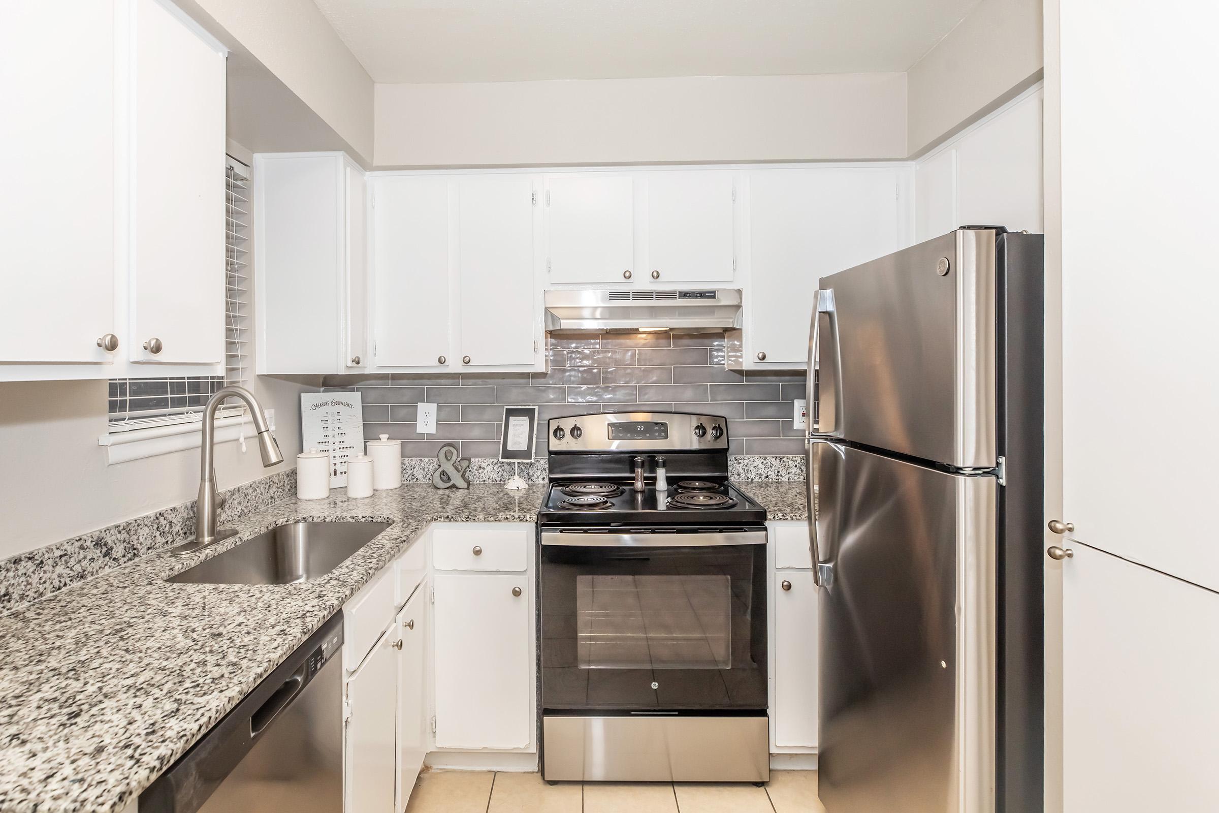 A modern kitchen with white cabinets, gray granite countertops, and stainless steel appliances. The kitchen features a silver refrigerator, a black oven and stovetop, and a sink with a faucet. Natural light filters in through a window, illuminating a tidy countertop with dishware and a decorative item.