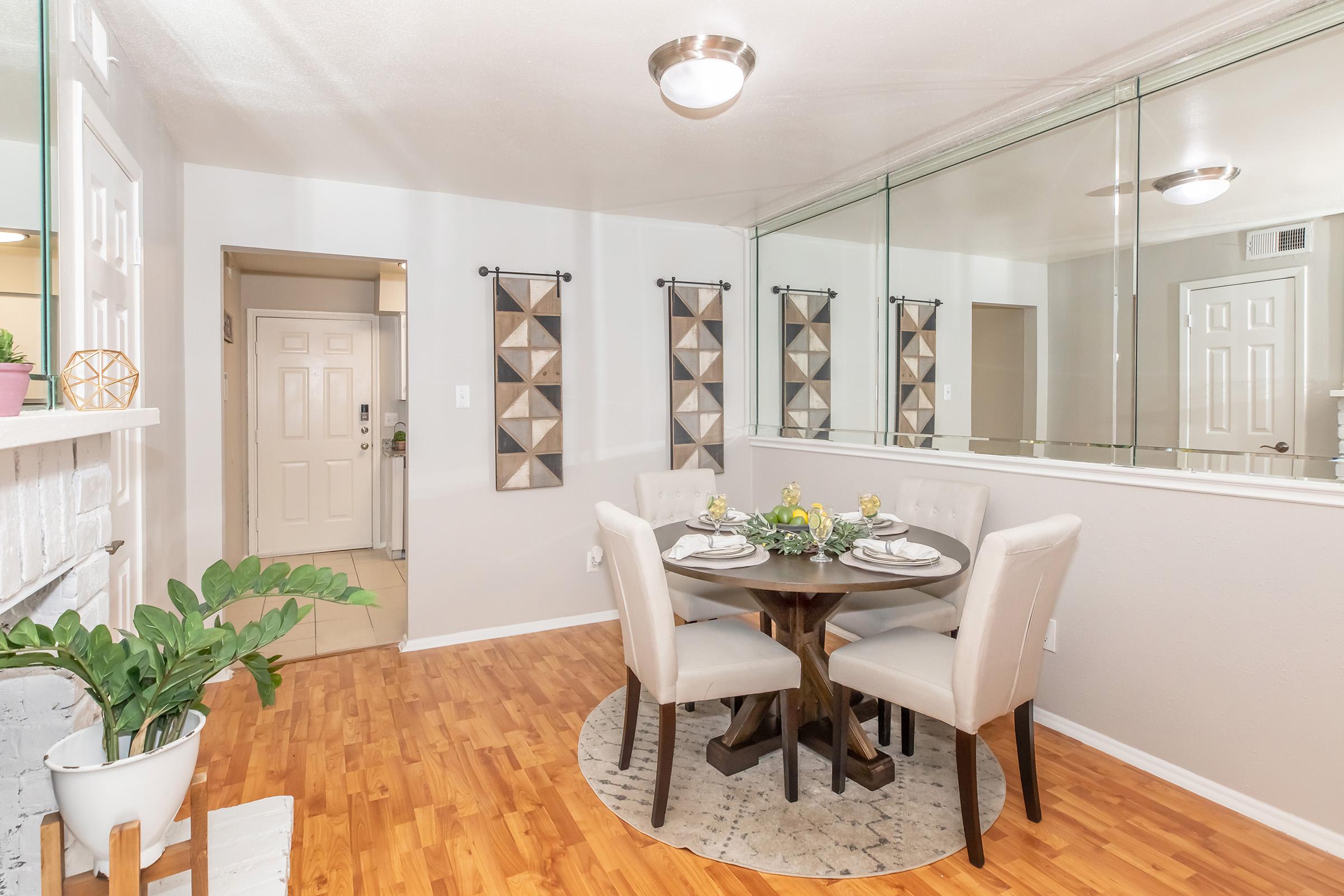 A cozy dining area featuring a round table with four upholstered chairs, set with plates and glasses. The wall behind has decorative mirrors, and two geometric-patterned wall hangings. A small indoor plant is placed beside the table, and a doorway can be seen leading to another room. The flooring is wooden, enhancing the warm ambiance.