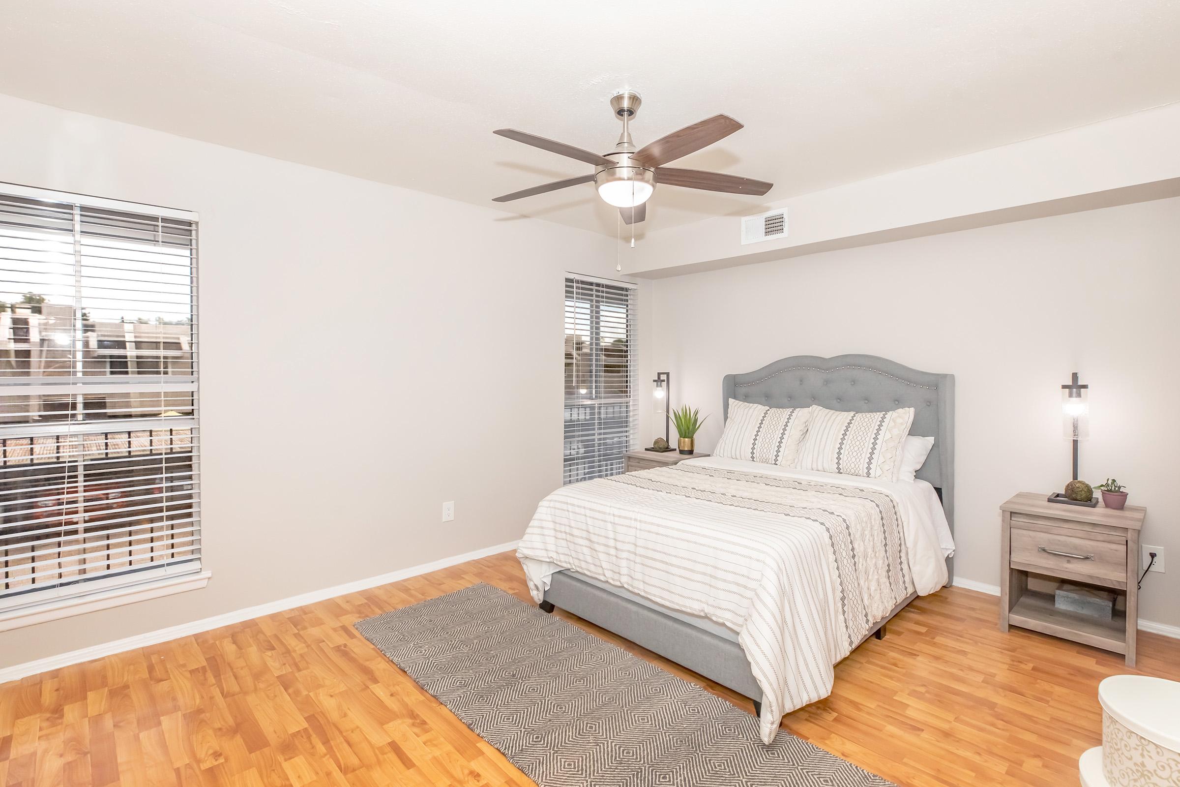 A cozy bedroom featuring a light gray bed with striped bedding, a wooden nightstand, and a ceiling fan. Natural light streams in through large windows with blinds, illuminating the room's modern decor and warm wooden flooring. A small plant adds a touch of greenery to the space.