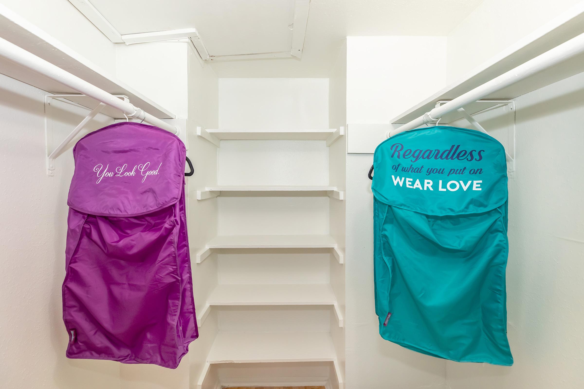 Two garment bags hanging in a closet. One bag is purple with the text "You Look Good," and the other is teal with "Regardless of what you put on WEAR LOVE." There are empty white shelves in the background.