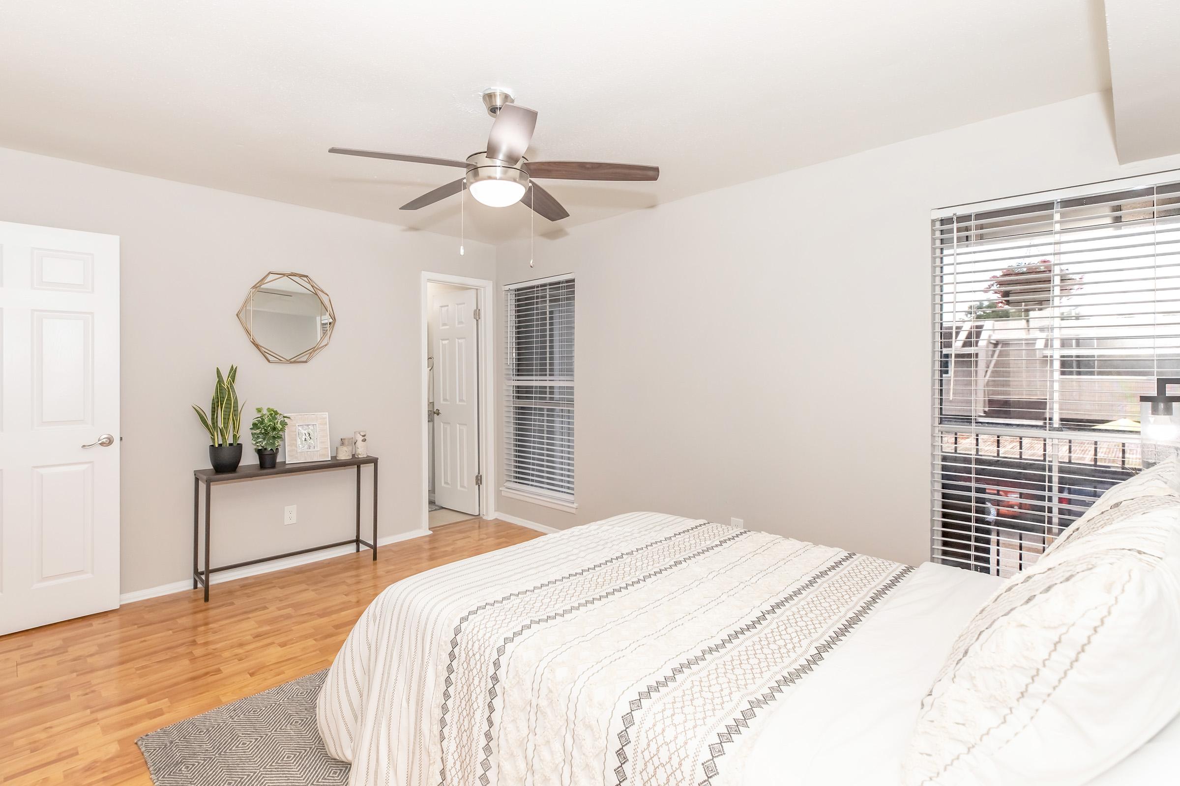 Bright and modern bedroom featuring a bed with light-colored linens, a ceiling fan, and a wooden floor. There is a small table with a plant next to the bed, and a large window with blinds allows natural light to fill the space. The room has neutral wall colors and a simple decor style.