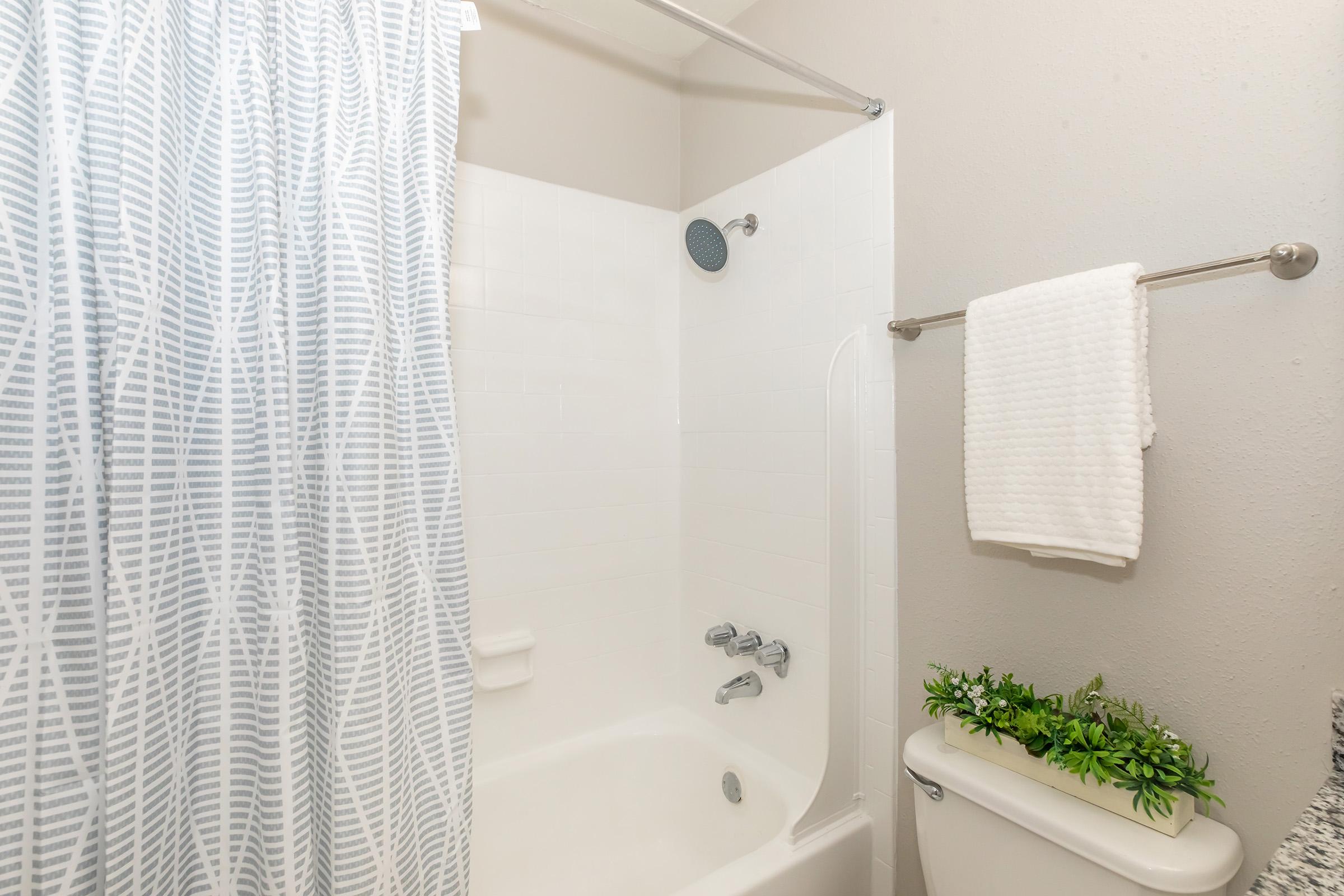 A clean, modern bathroom featuring a shower with a white and gray patterned curtain, a white bathtub, and a chrome showerhead. A towel is hanging on a rod next to a small potted plant on the edge of the toilet, which is also white. The walls are a light neutral color.