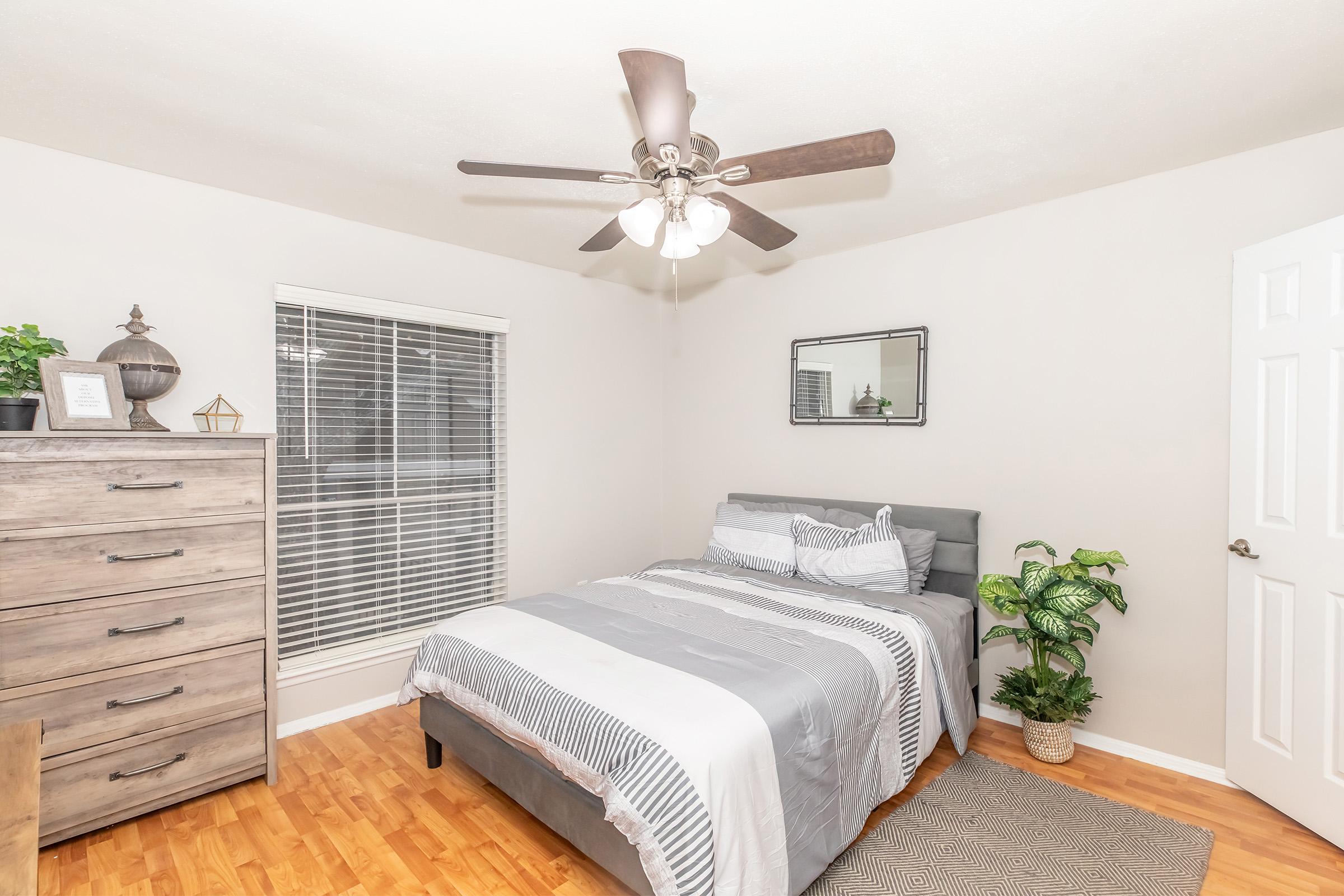 A cozy bedroom featuring a gray bed with striped bedding, a wooden dresser, a fan with lights, and a small plant. A window with blinds allows natural light, and a mirror is mounted on the wall. The floor is wooden, adding warmth to the room's neutral color palette.