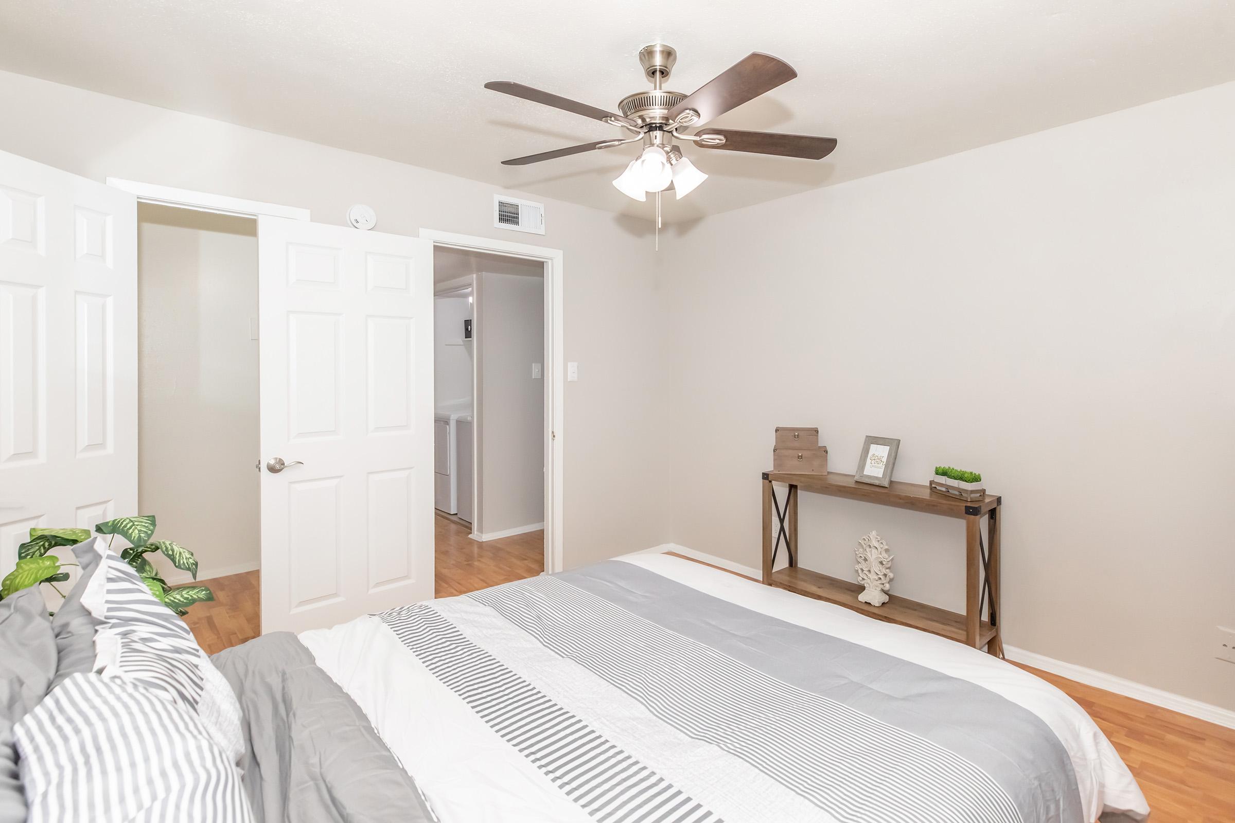 A cozy bedroom featuring a bed with gray and white bedding, a ceiling fan, and a small wooden table with decorative items. Two white doors open to adjacent spaces, with hardwood flooring and soft, neutral-colored walls. Green plants add a touch of nature to the room.