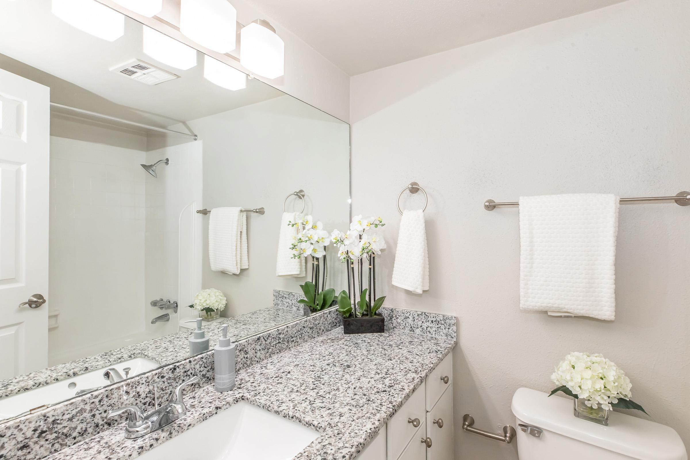 A modern bathroom featuring a light-colored granite countertop with a sink, a large mirror, and a silver towel rack with two white towels. Orchids in a planter sit on the counter, and a small bouquet of white flowers is placed next to the sink. Soft lighting from overhead fixtures adds to the clean, elegant atmosphere.