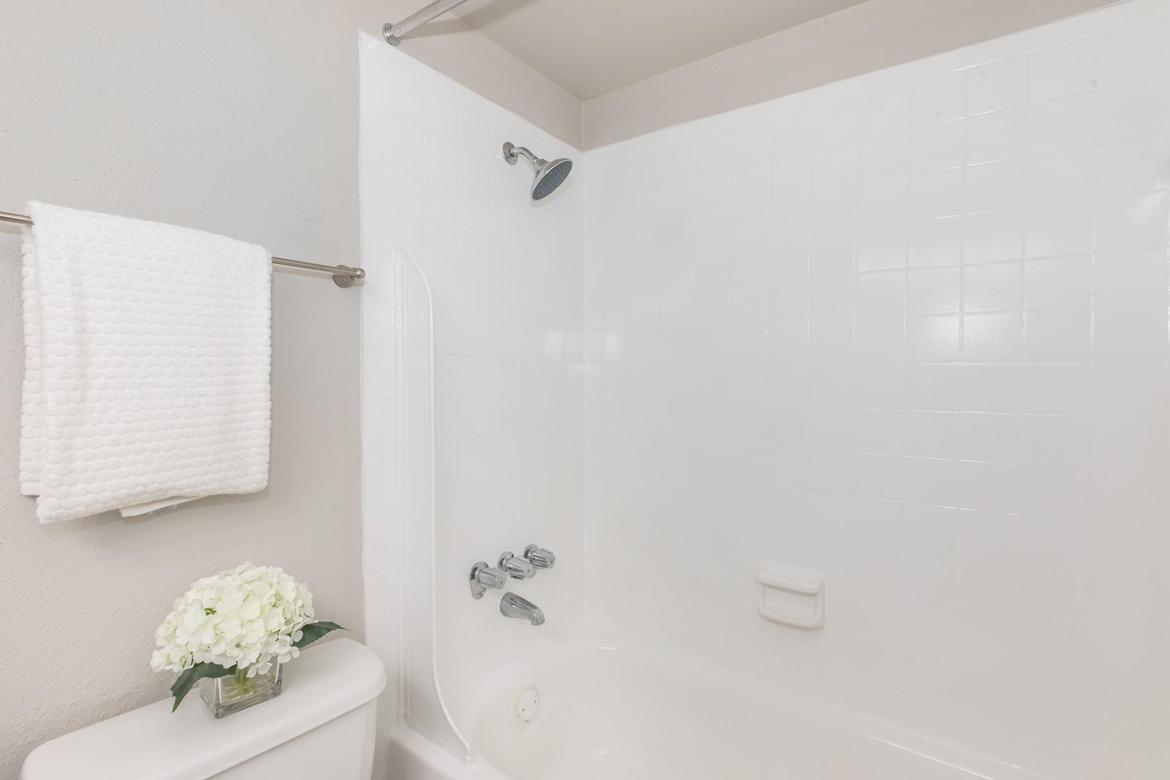 A clean, modern bathroom featuring a white tiled shower with a showerhead, a white towel hanging on a bar, and a small vase of white flowers on a countertop next to a toilet. The overall aesthetic is bright and minimalistic.