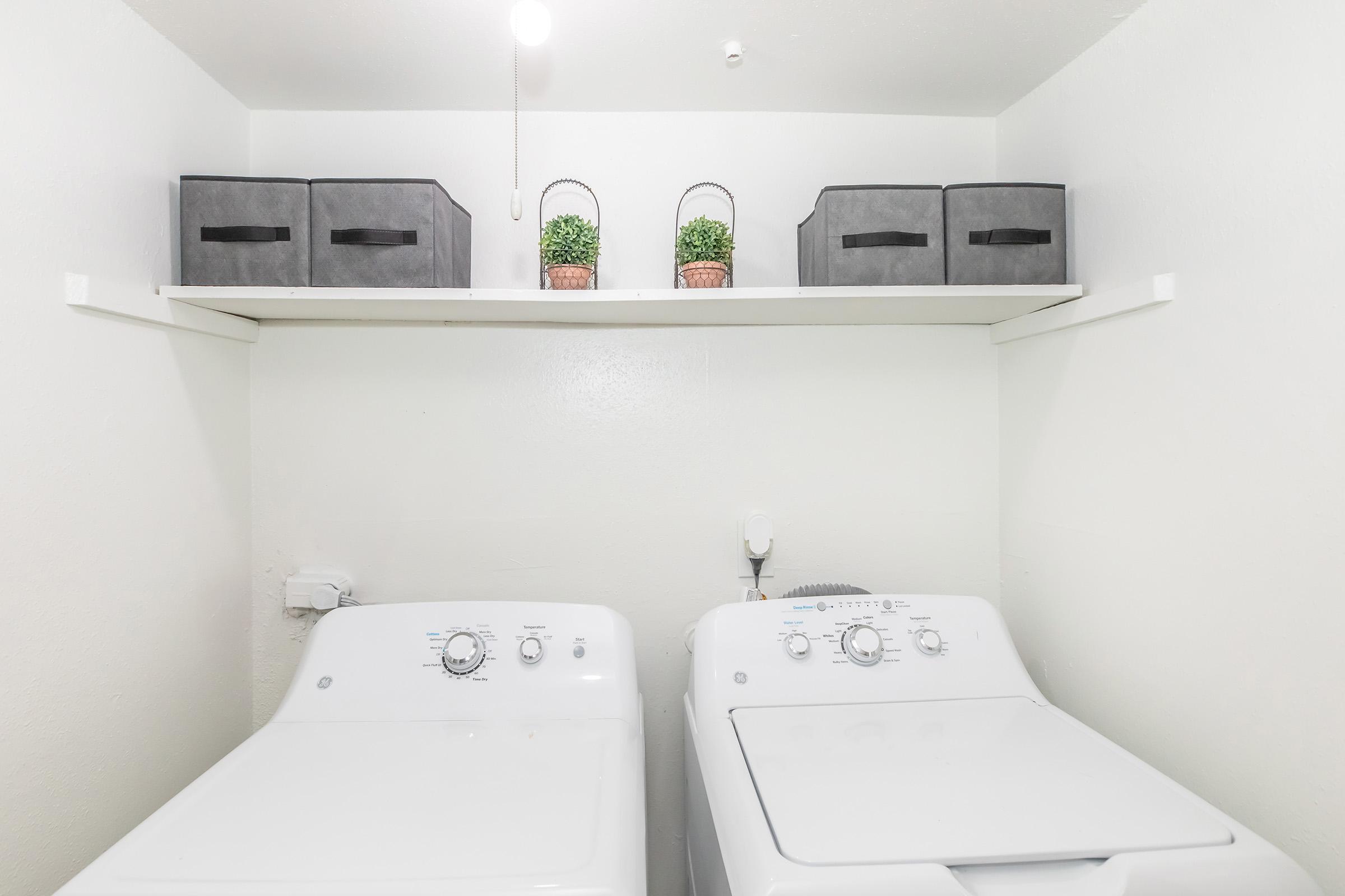 A clean laundry room featuring a white washing machine and dryer side by side. Above them, a shelf holds two grey storage boxes and two small potted plants, adding a touch of greenery to the minimalistic space. The walls are painted white, creating a bright and airy atmosphere.