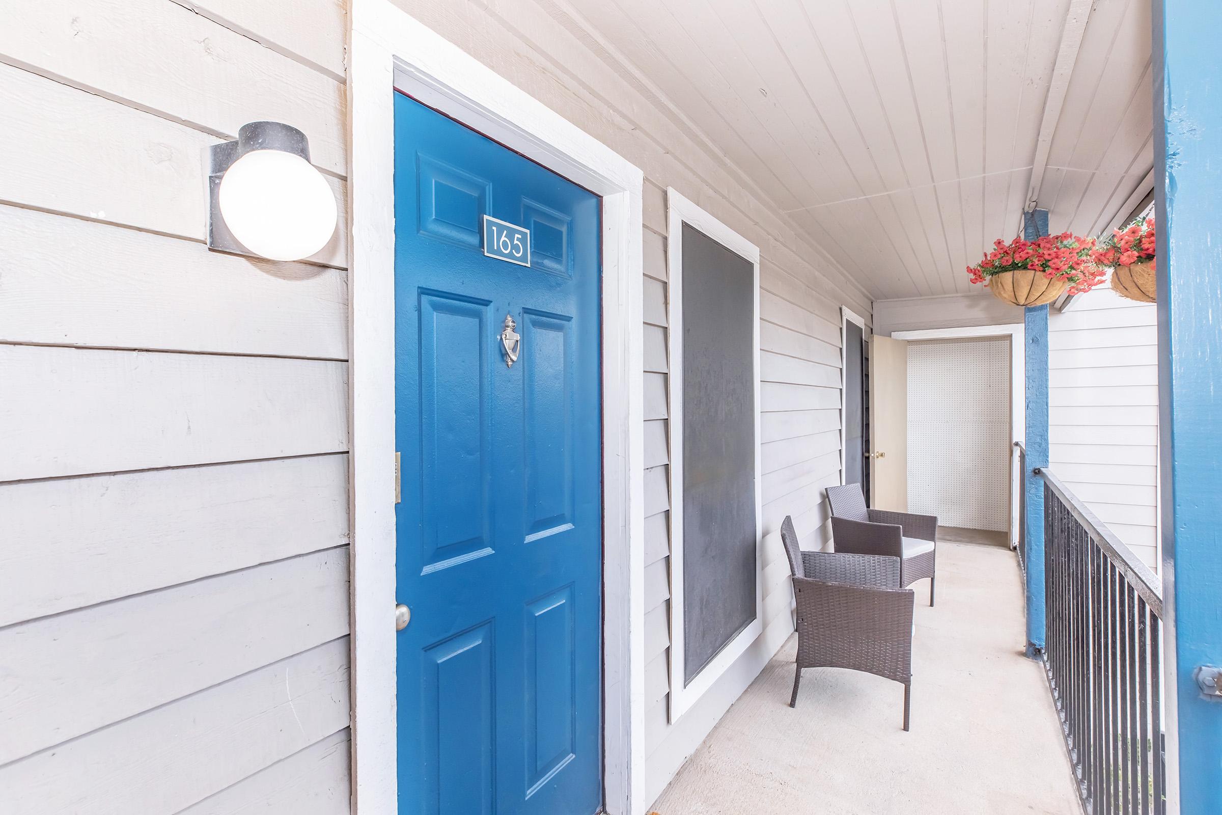A view of a hallway with a bright blue door marked "165." Two wicker chairs are placed near the door, and a hanging flower basket adds a touch of color. The walls are light gray, and there is a large black panel beside the door. The area is well-lit, creating a welcoming atmosphere.