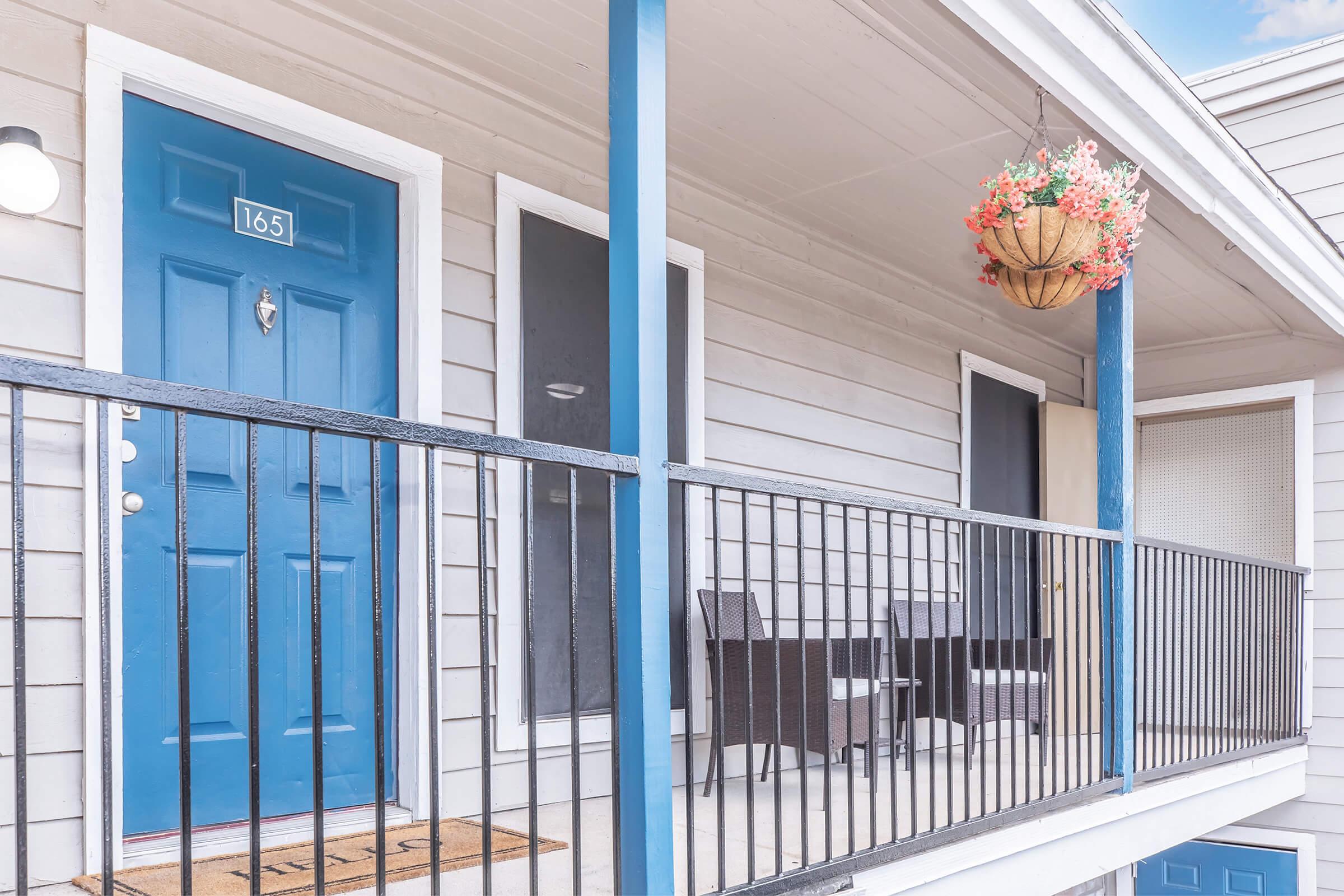 A well-lit exterior view of a balcony with a blue door labeled "165." The balcony features a black railing, two wicker chairs, and a hanging flower basket. The building has light gray siding, two window frames, and a welcoming atmosphere with a doormat in front of the door.