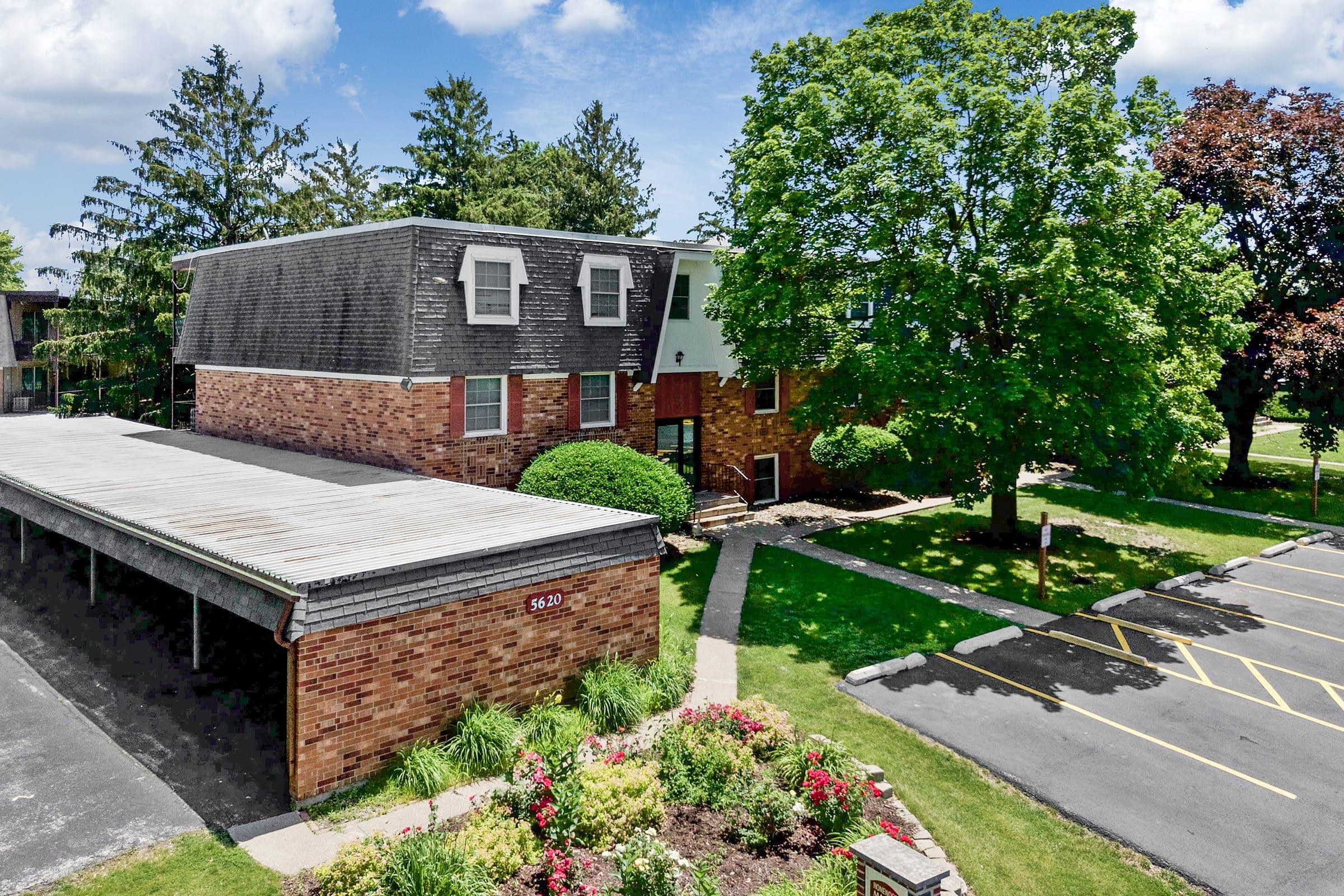Aerial view of a landscaped apartment complex featuring a brick building with green shrubs and trees surrounding it. A covered parking area is visible on the left, and a pathway leads through flower beds in the foreground. The sky is partly cloudy, adding to the pleasant atmosphere.