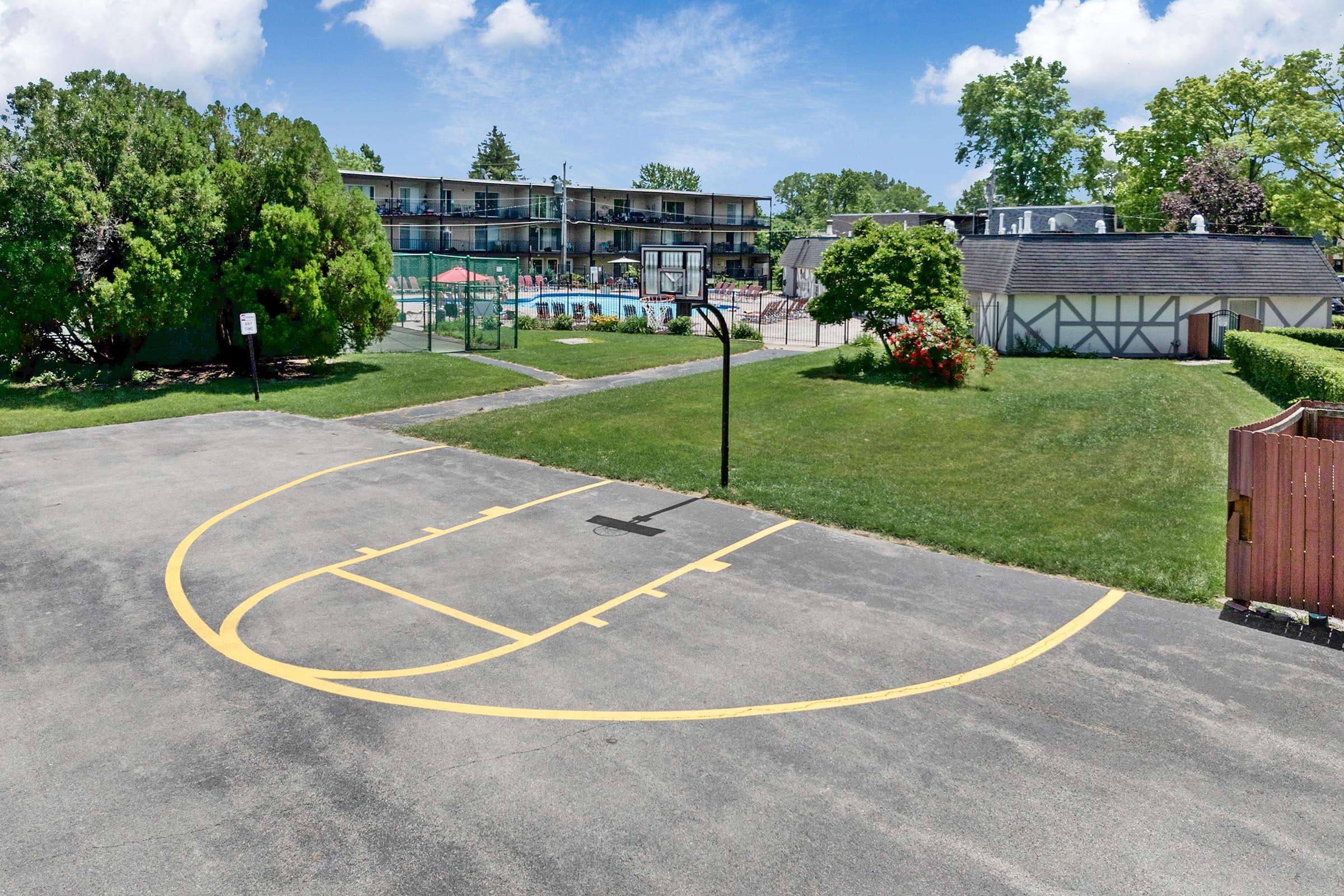 A basketball court marked with yellow lines, located outdoors. In the background, there's a motel with multiple units and a swimming pool. Surrounding the area are green trees and well-kept grass, creating a relaxed atmosphere.