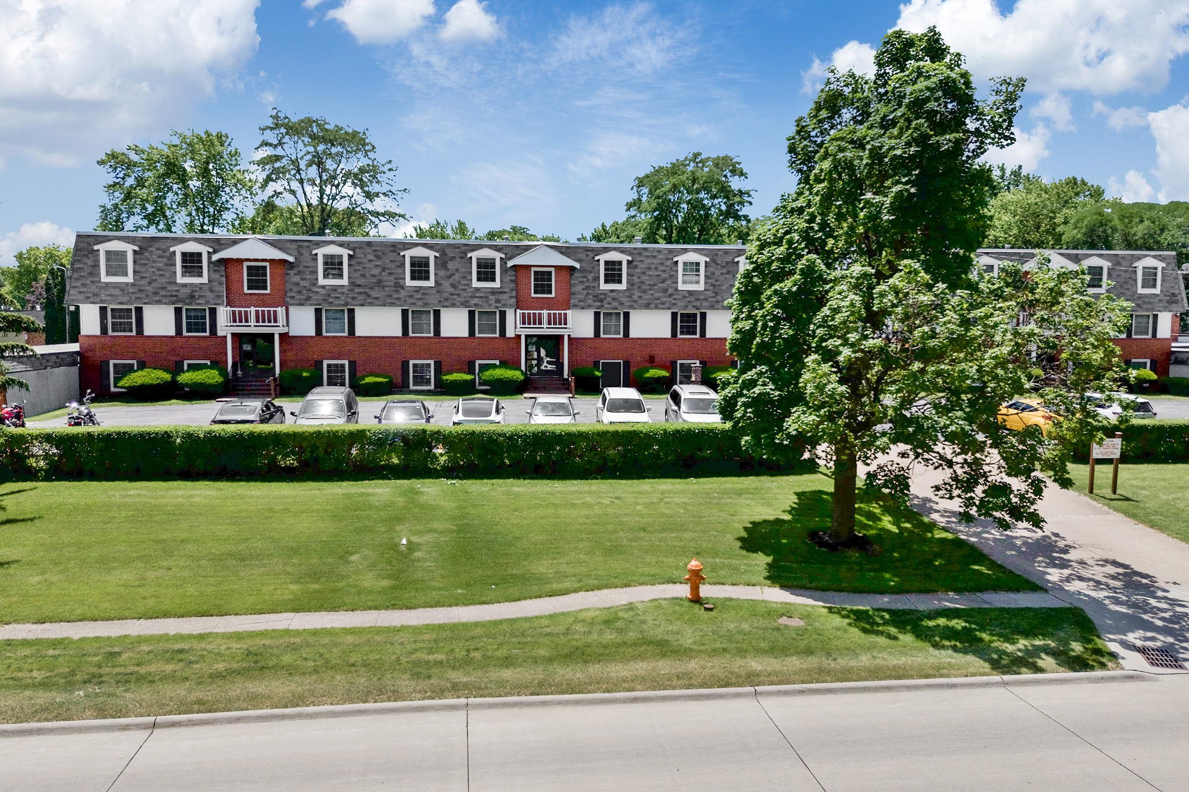 A row of two-story townhouses with gray roofs and brick exteriors, surrounded by well-maintained lawns and landscaping. Several parked cars line the driveway in front of the homes, and a large tree stands prominently to the right. Bright blue sky with scattered clouds overhead.