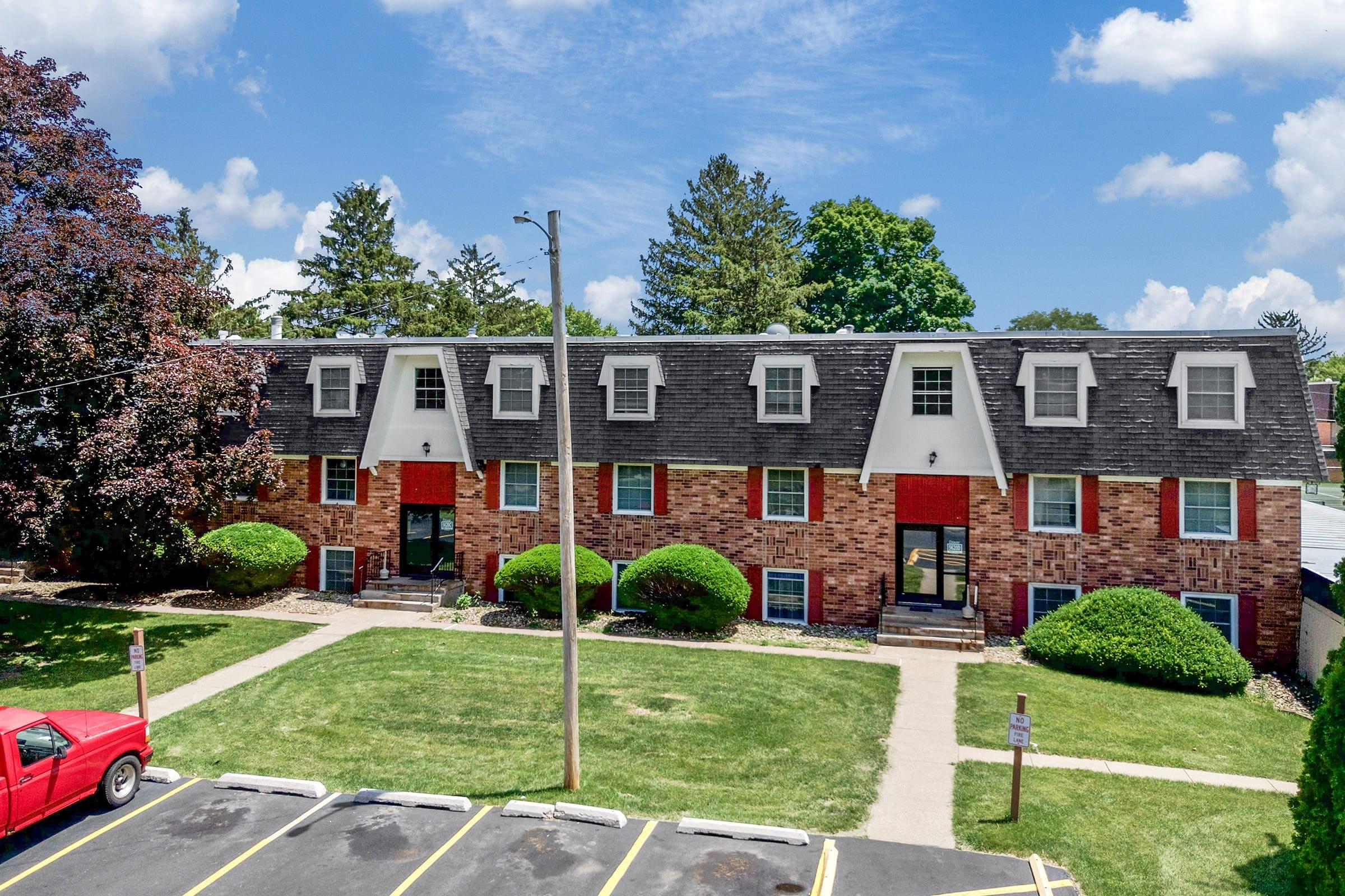 A brick apartment building with multiple units, featuring a sloped roof and large windows, surrounded by well-maintained green lawns and shrubs. There is a red car parked in a lot in front of the building, with clear blue skies and a few clouds in the background.