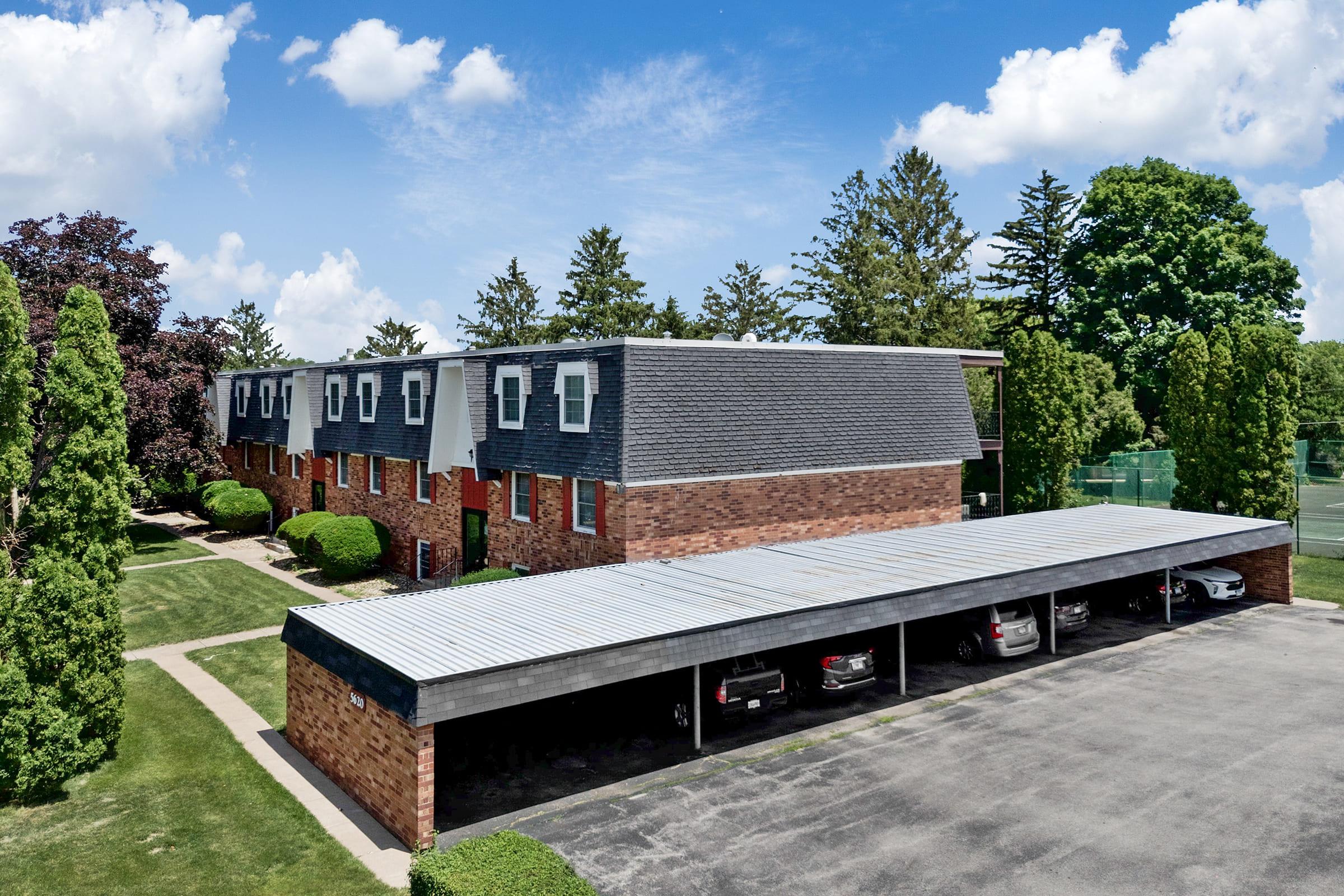 Aerial view of a brick apartment building with a modern facade, surrounded by greenery. Below, a covered parking area is visible with several parked cars. The sky is clear with few clouds, and the landscape features trees and manicured lawns.