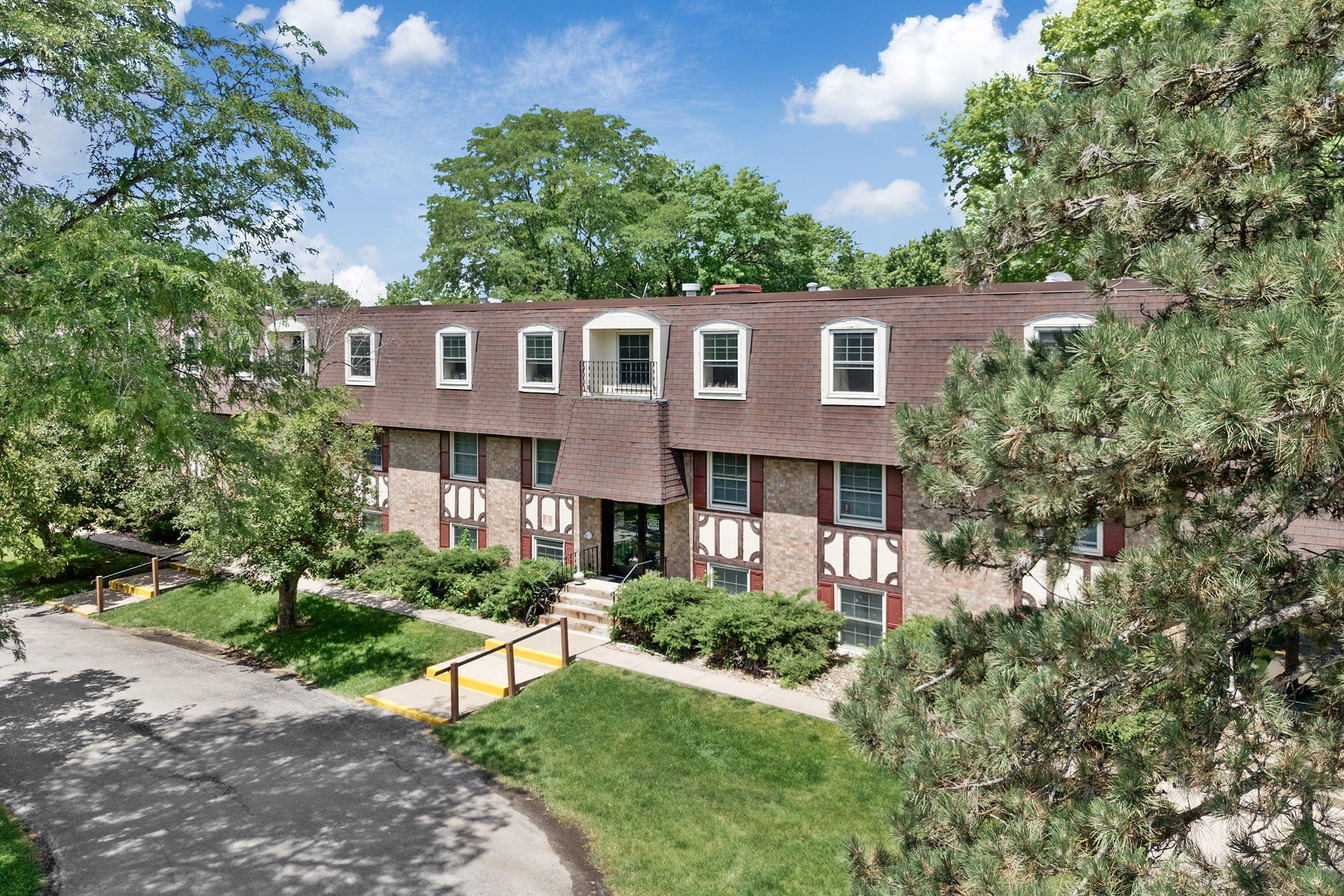 Exterior view of a multi-story residential building surrounded by greenery, with a blue sky and clouds in the background. The building features a mix of brick and siding, multiple windows, and a central entrance, alongside a pathway and landscaped areas.