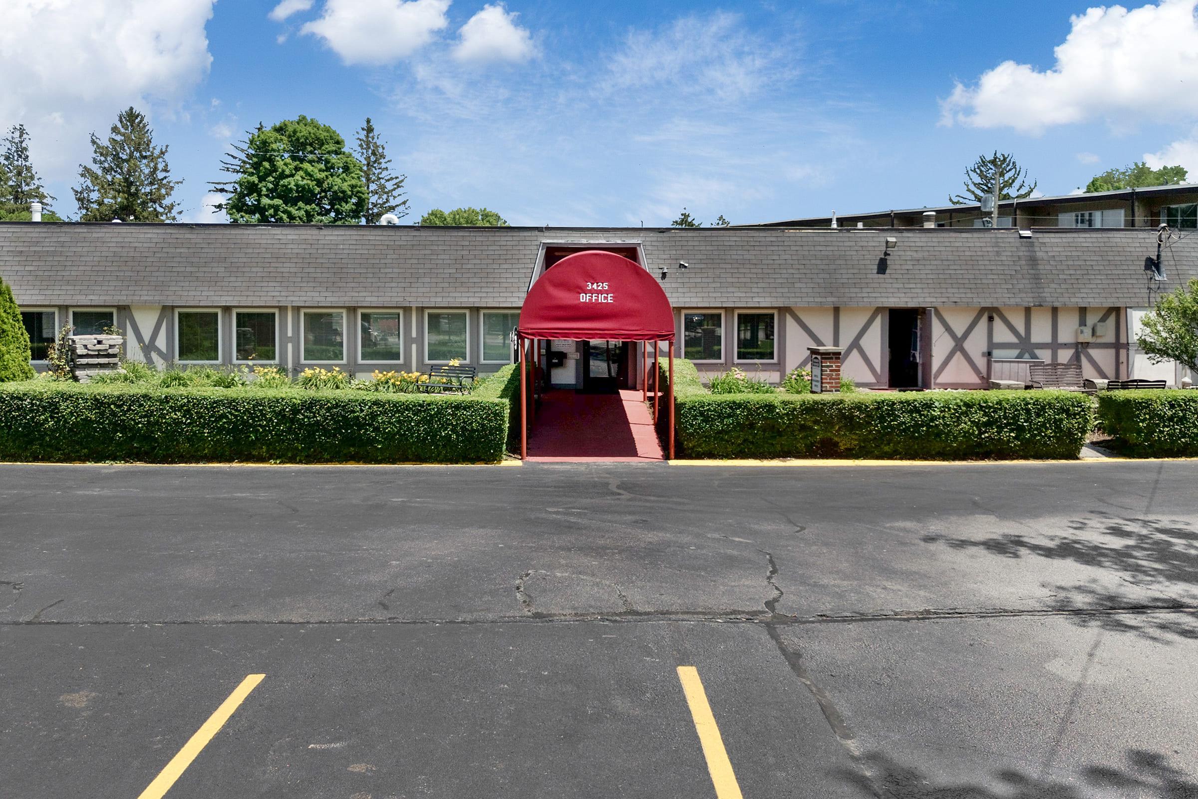 A single-story building with a red awning labeled "Lodge Office" at the entrance. The building features a landscaped front with green shrubs and flowerbeds, surrounded by a paved parking lot. In the background, there are trees and a blue sky with scattered clouds.